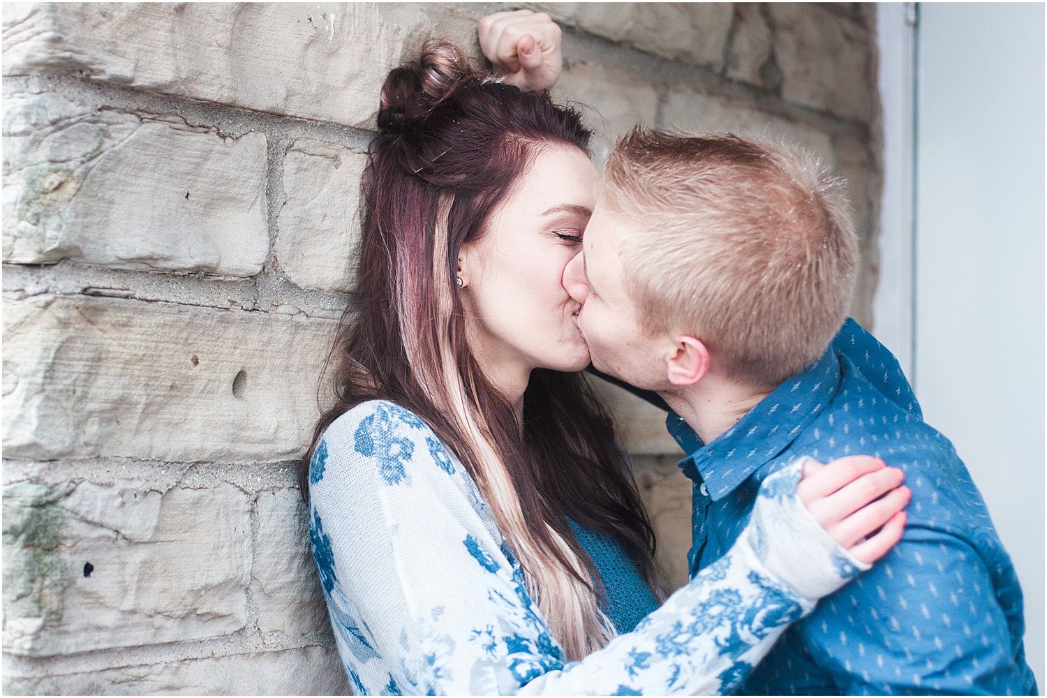 A couple shares a kiss against a textured stone wall during their winter photoshoot.