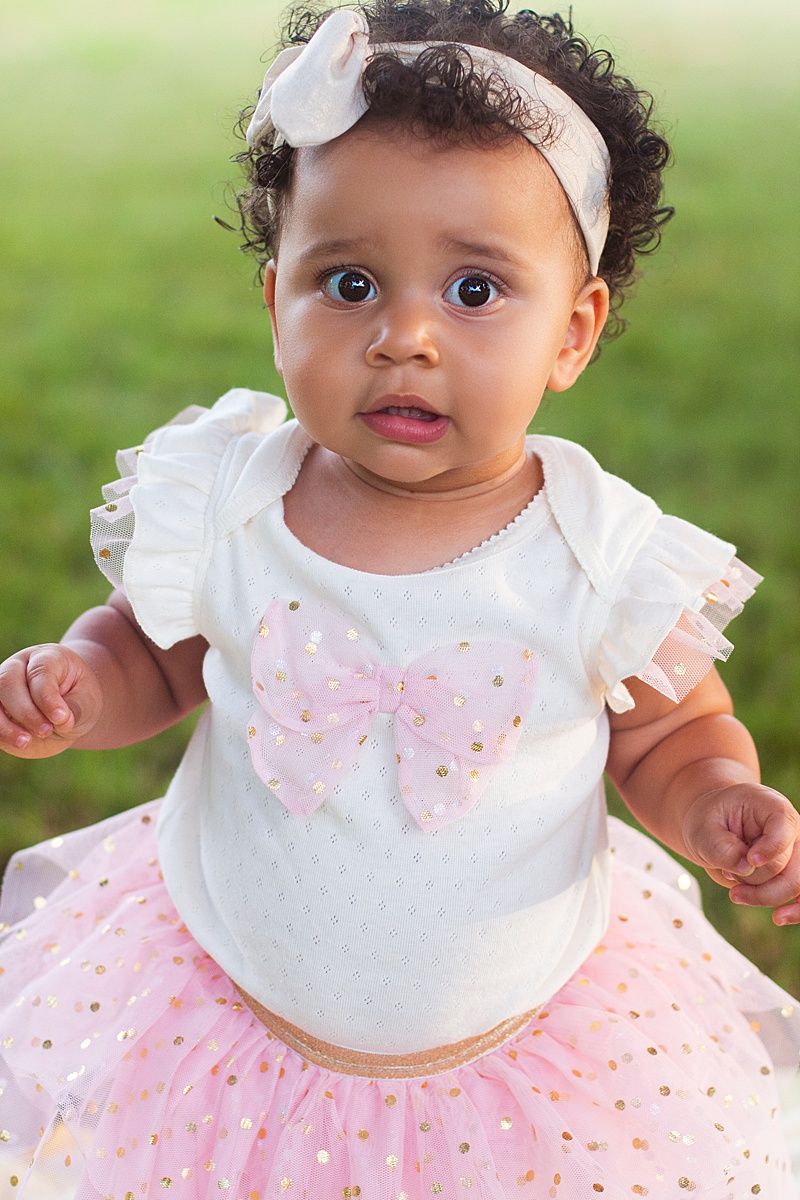 Baby in tutu dress with butterfly design and white headbow standing against green background.