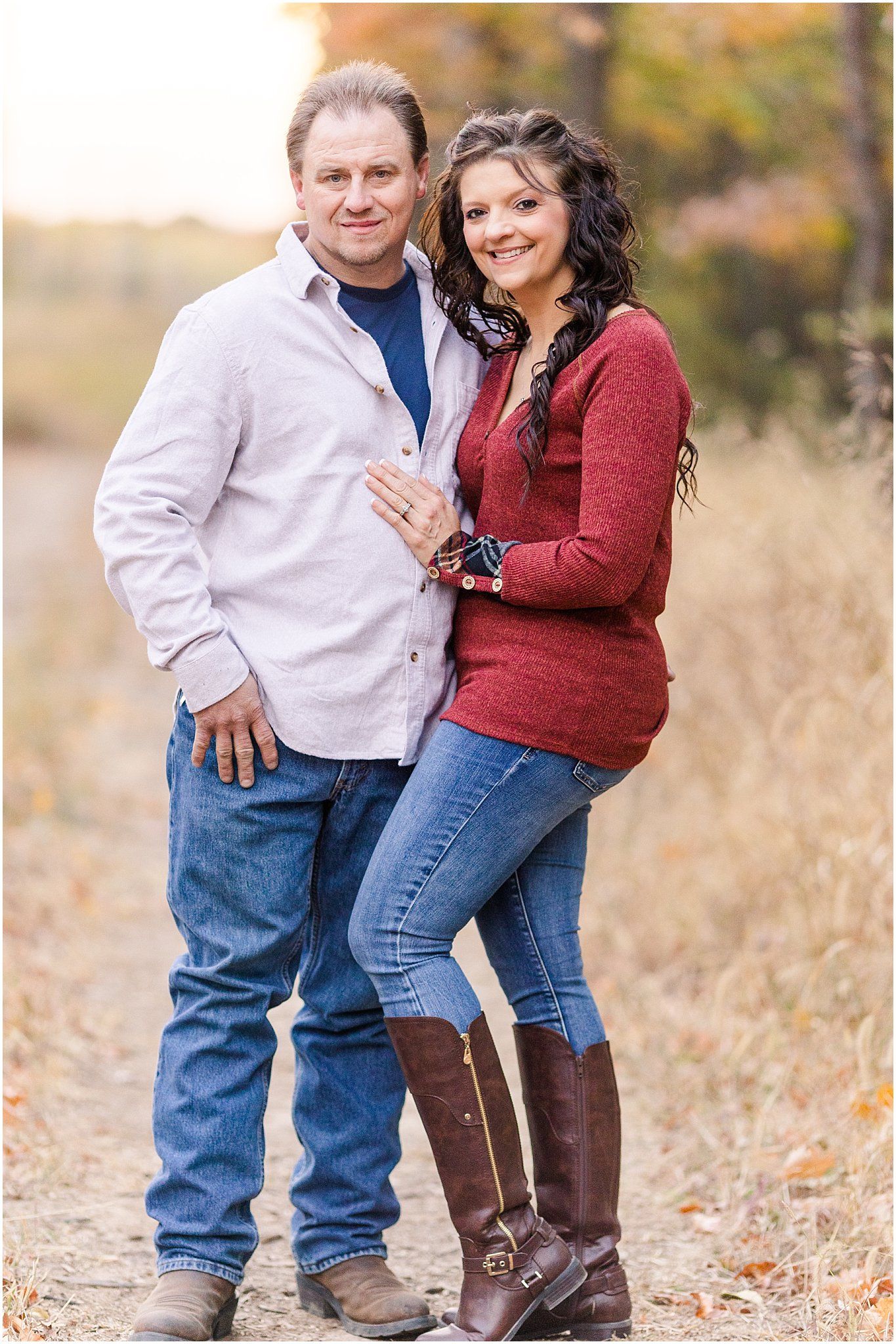 Couple in casual fall attire pose together outdoors with autumn foliage in background.