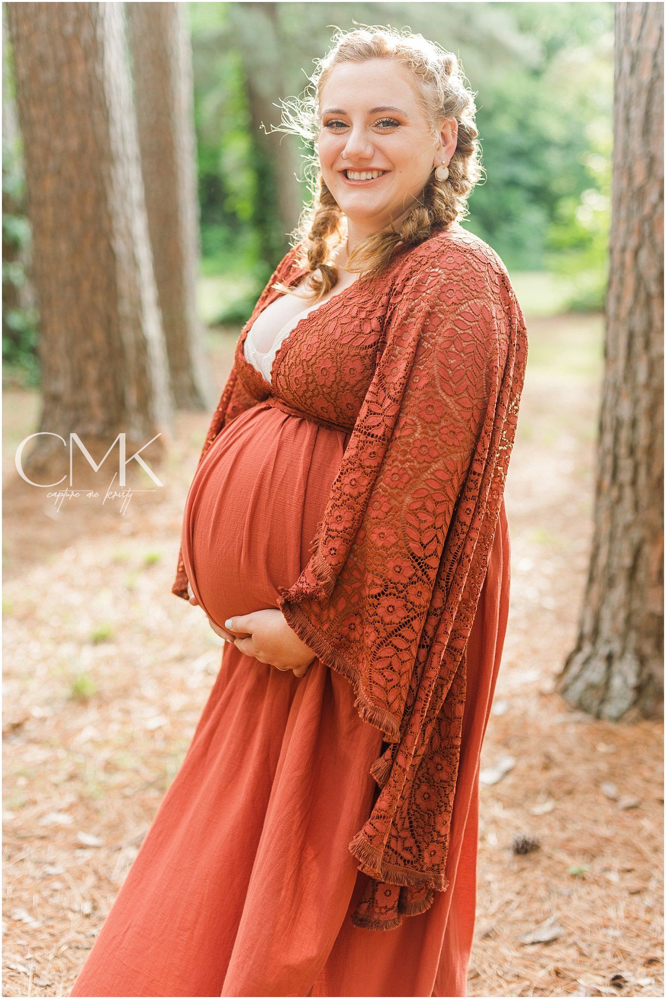 Pregnant woman in rustic dress smiles happily during outdoor maternity session.