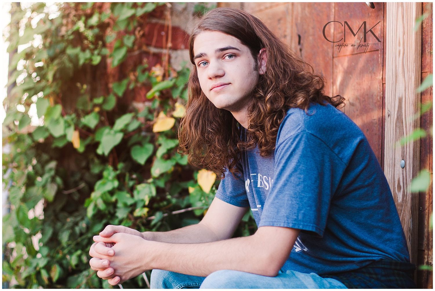 Person in blue t-shirt sitting beside climbing vines and brick wall.