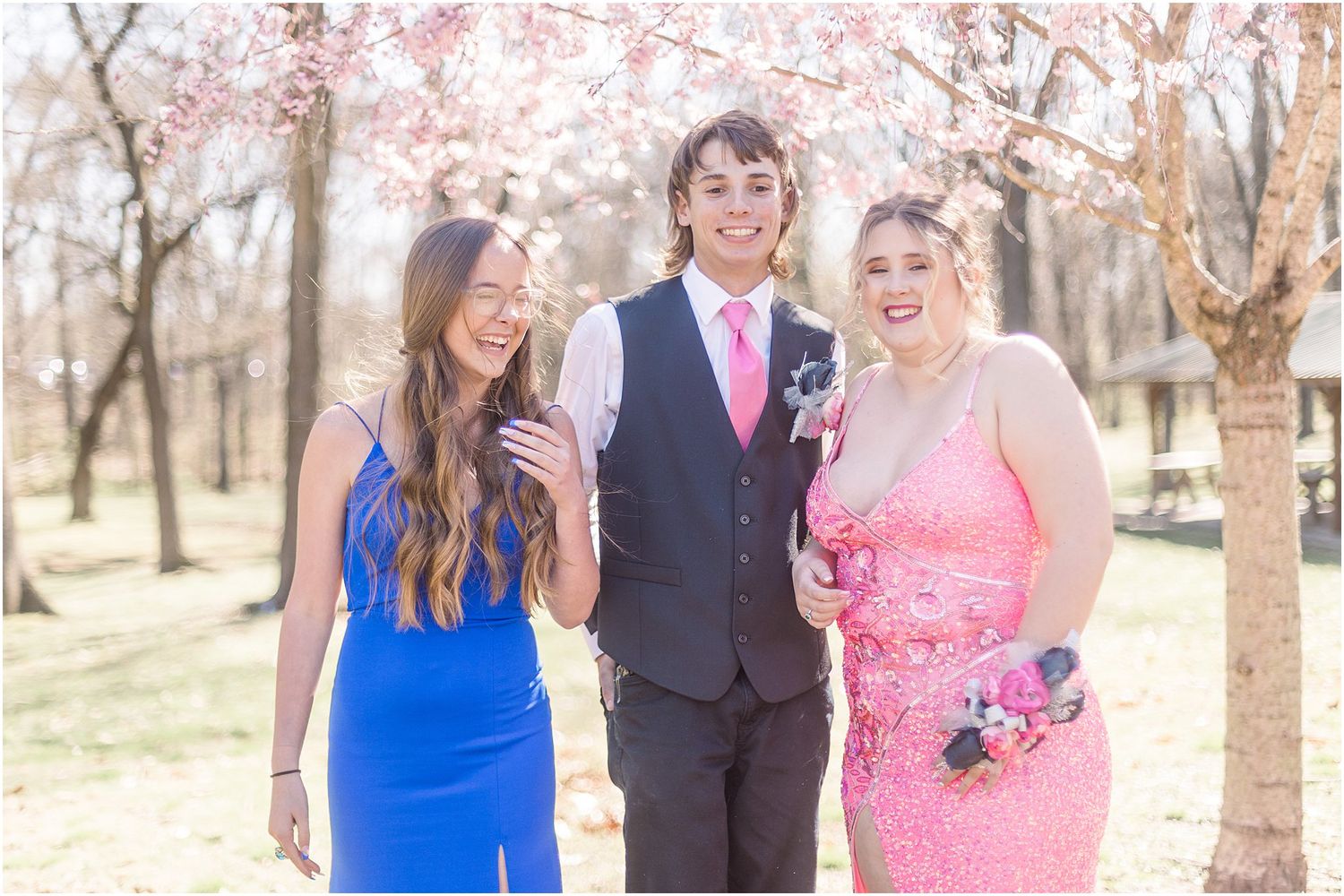 Three friends share a candid laugh while posing in their prom outfits under flowering trees.
