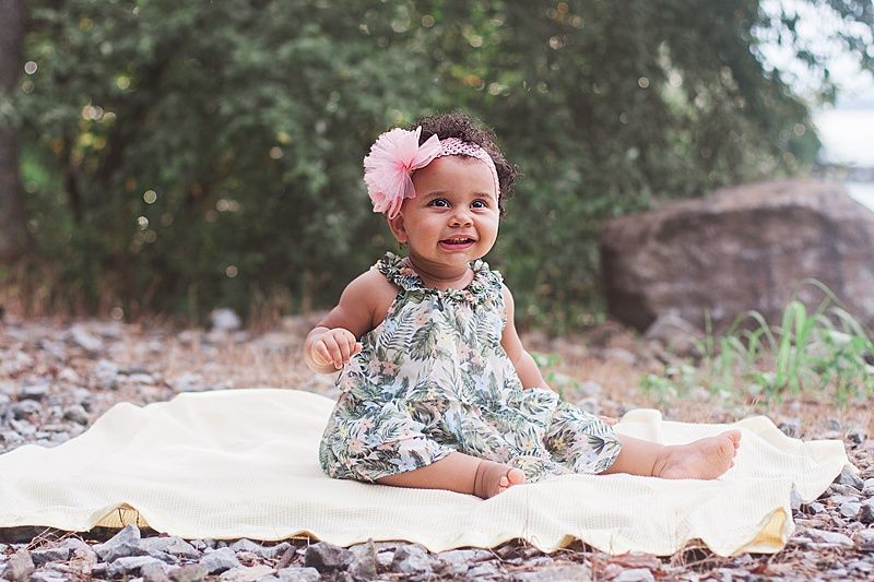 Baby in floral dress and pink bow sitting on white blanket outdoors.