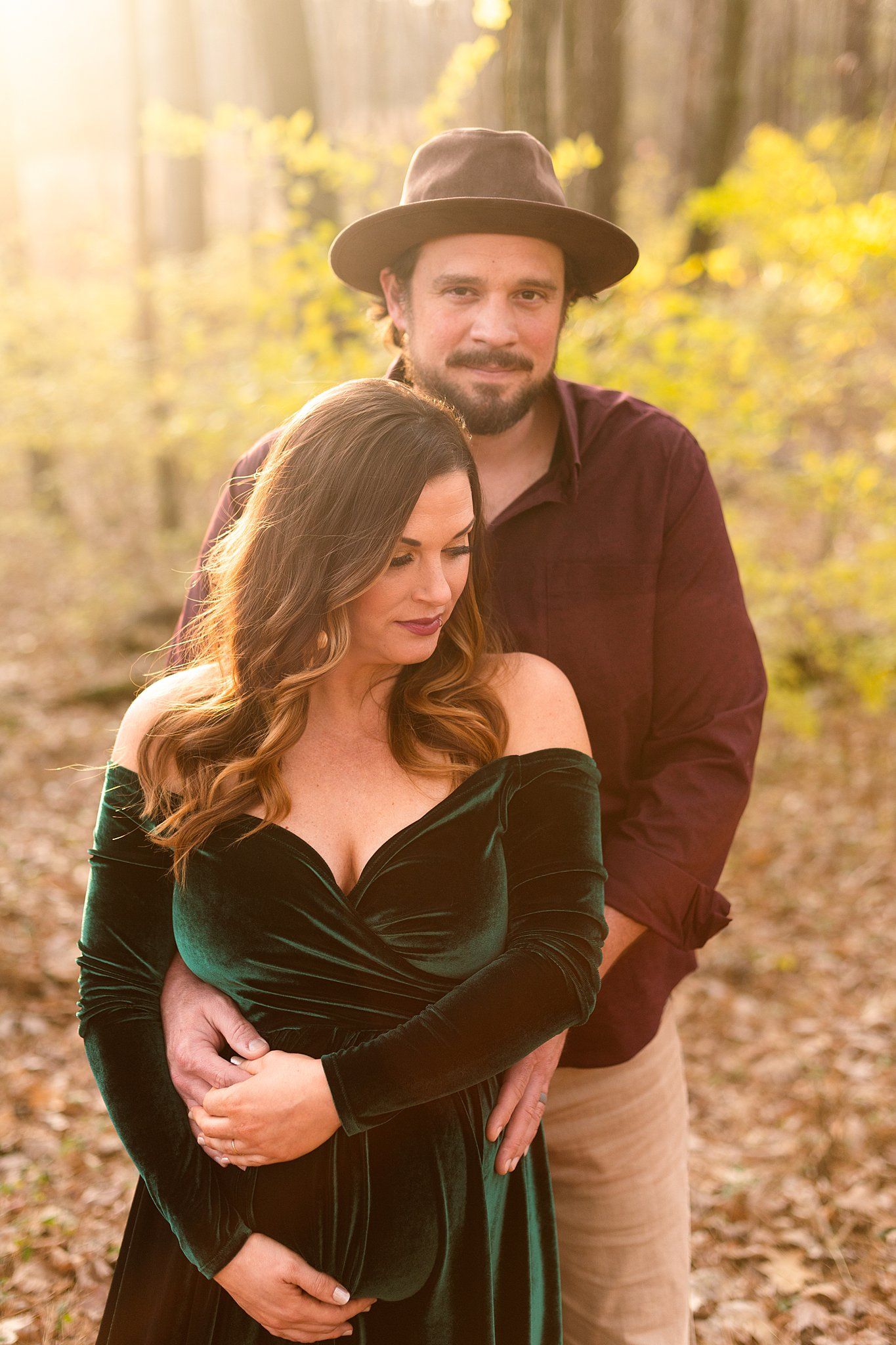 A romantic couple embraces in an autumn forest setting with sunlit trees, wearing a brown hat and emerald velvet dress.