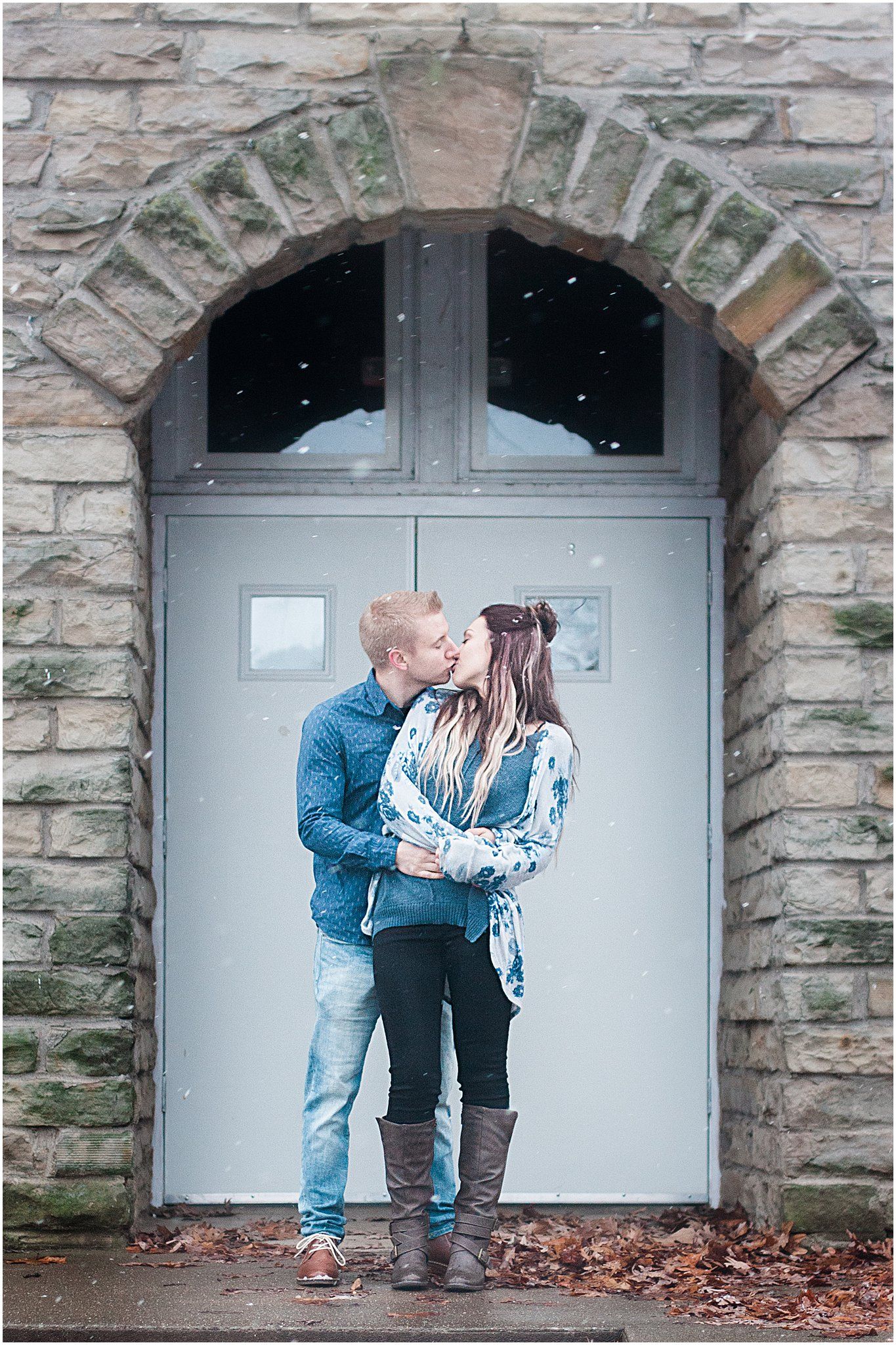 A couple shares a romantic moment under an arched stone doorway during a light snowfall.