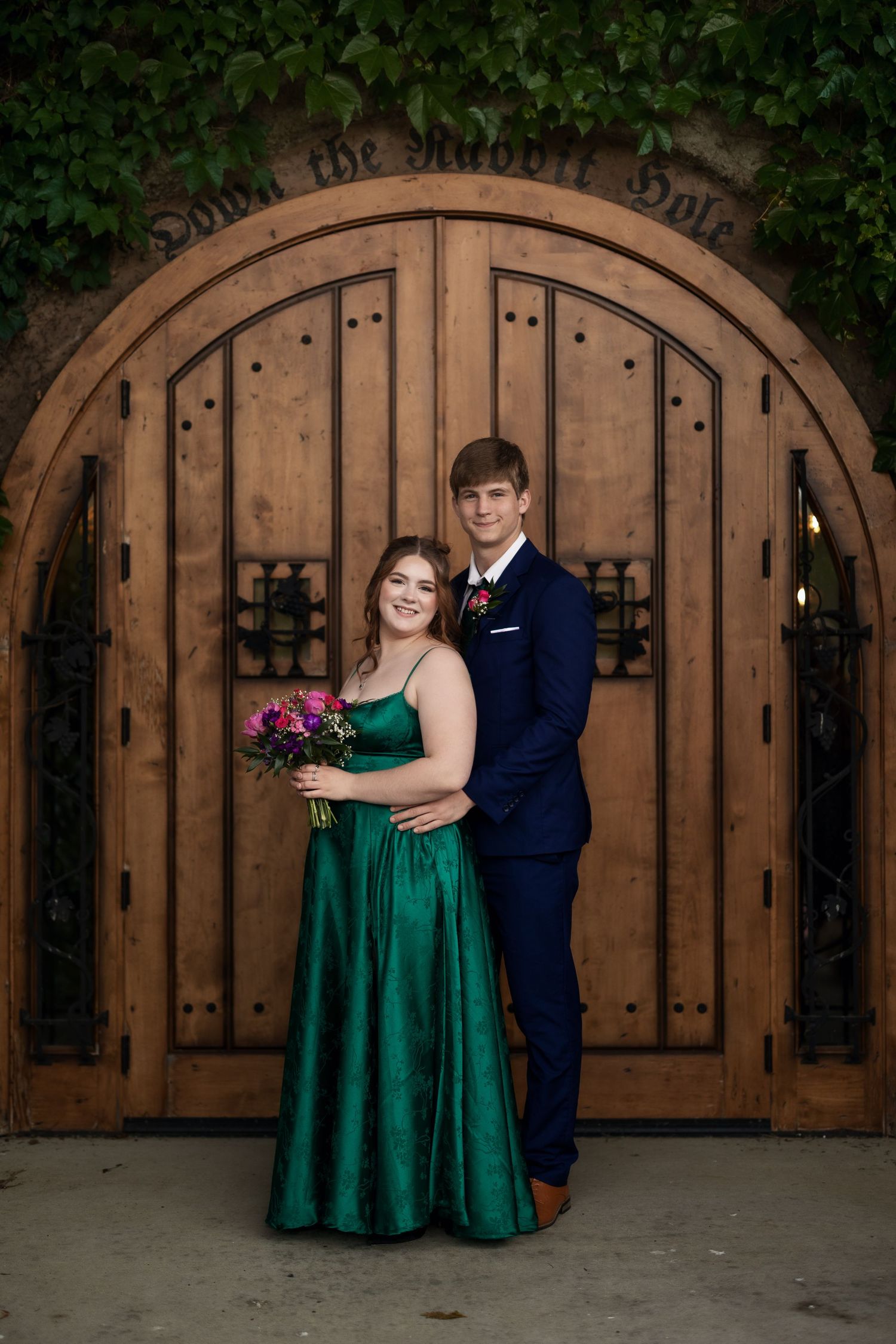 A couple in prom attire poses before a wooden arched door, she in emerald gown, he in navy suit with boutonniere.