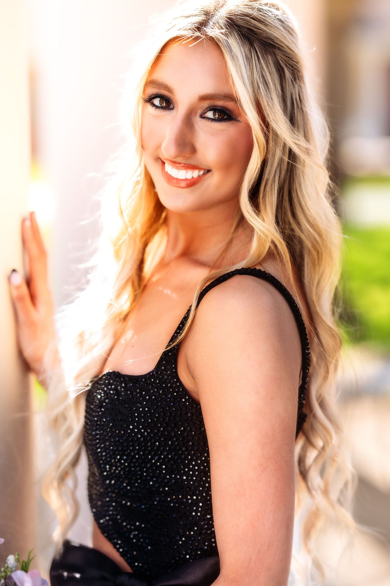 A person in a sparkly black formal dress poses outdoors against a blurred architectural background.