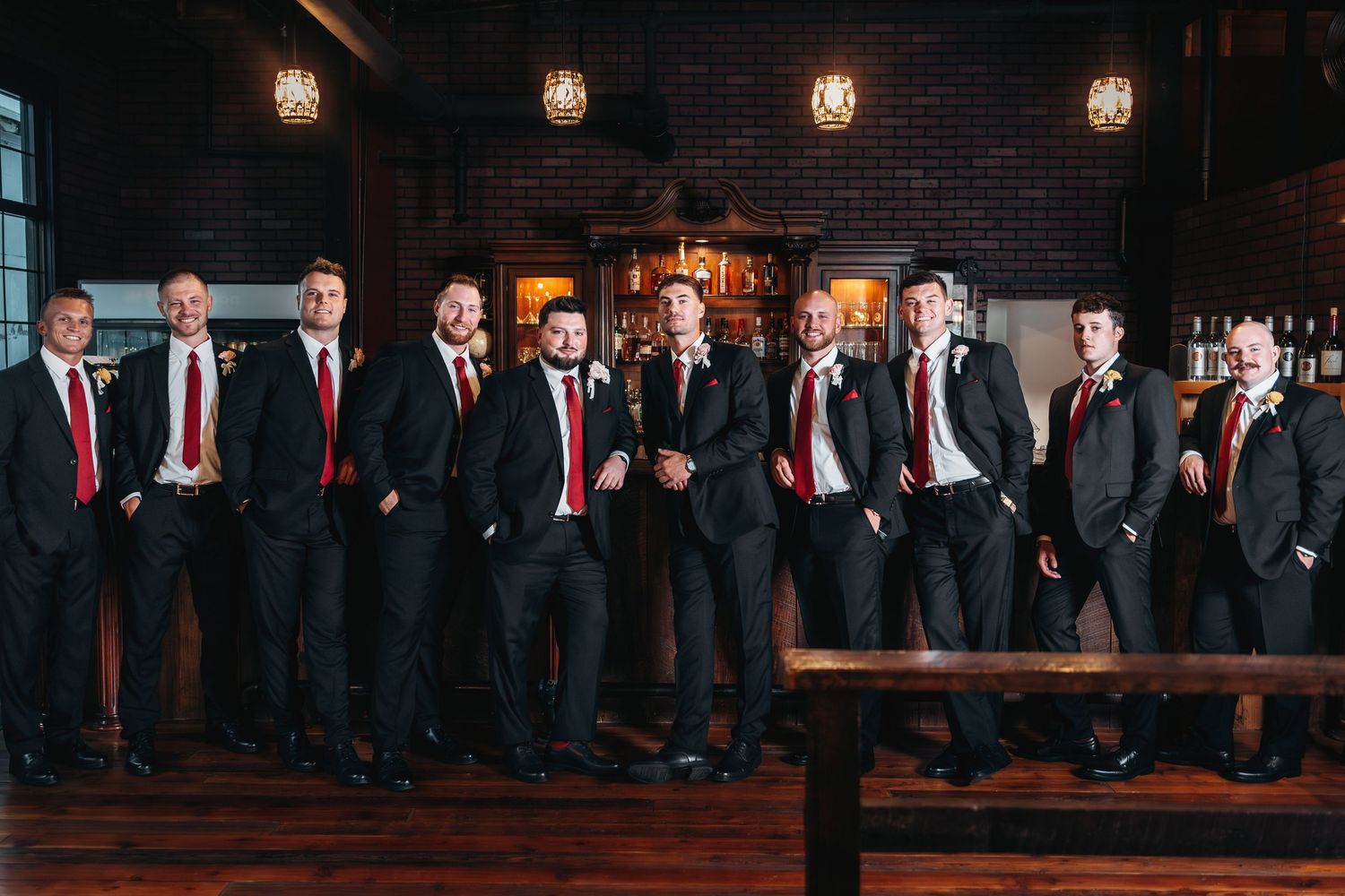Groomsmen in dark suits pose against a rustic brick wall with warm lighting.