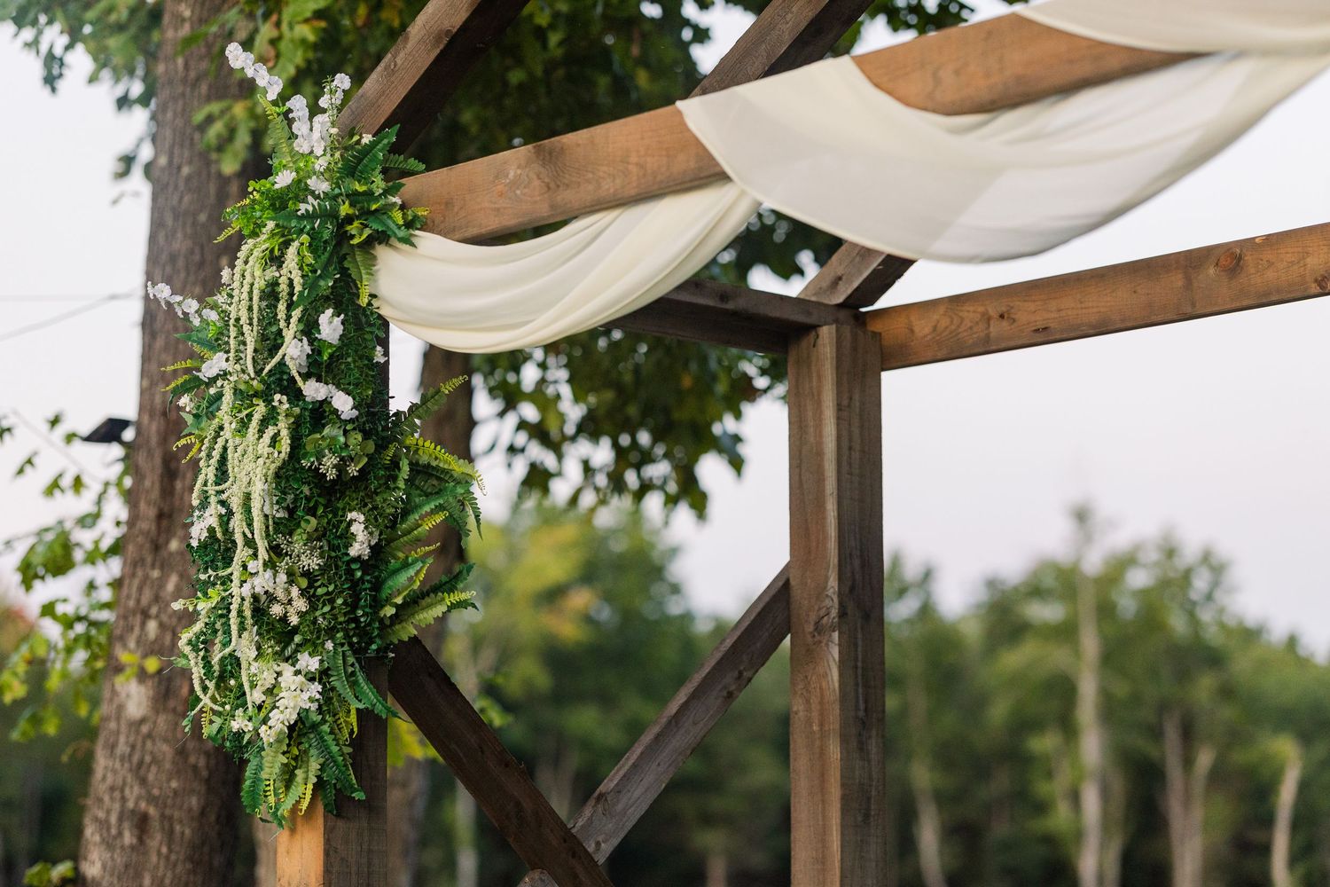 Wedding ceremony pergola decorated with white fabric and greenery against a natural backdrop.