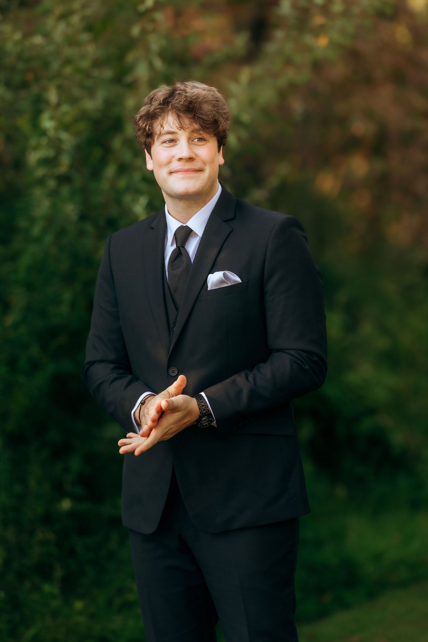 Groomsman in black suit poses playfully for formal wedding photos outdoors.