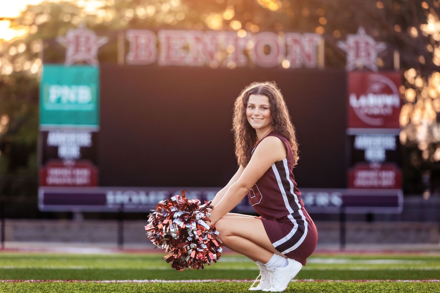 Cheerleader in maroon uniform poses with pom-poms on football field with Benton scoreboard backdrop.