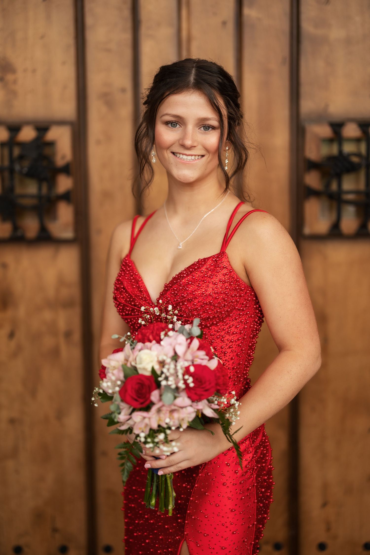 Smiling young woman in a red sequined gown holding a bouquet of roses and flowers, posing in front of wooden doors.