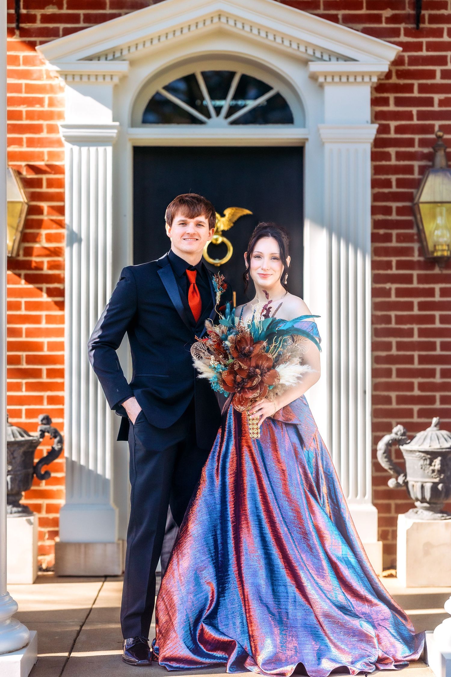Couple in formal attire poses outside a brick building with white columns and doorways.