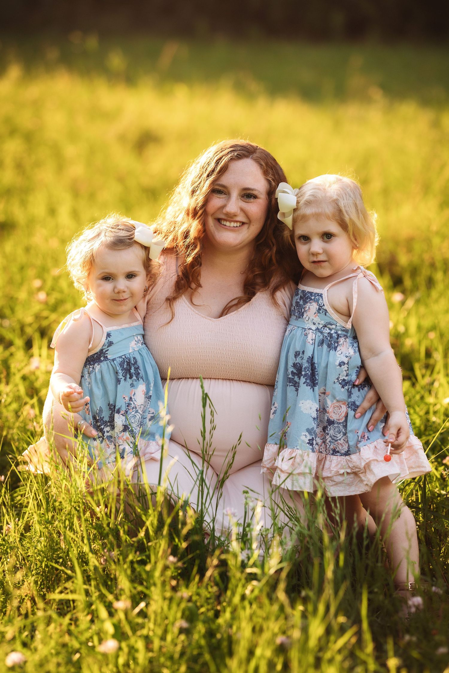 A maternity photo in a sunlit meadow shows matching blue floral dresses against golden grass at sunset.