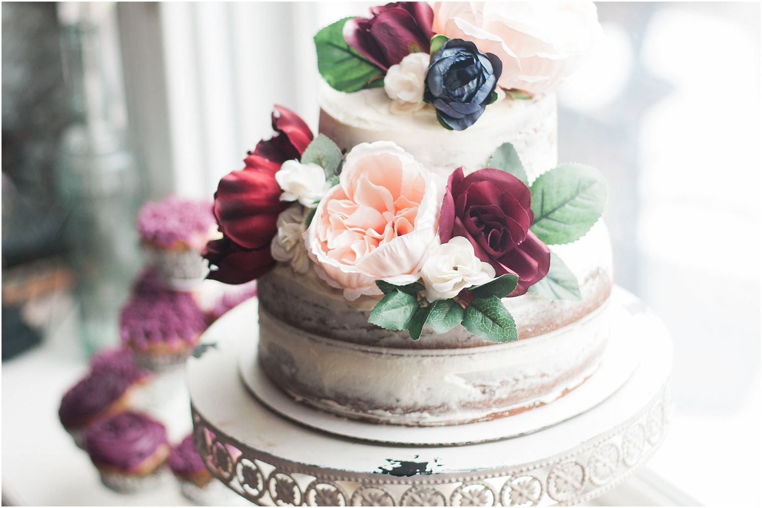 Semi-naked wedding cake decorated with burgundy and peach roses on an ornate white stand.
