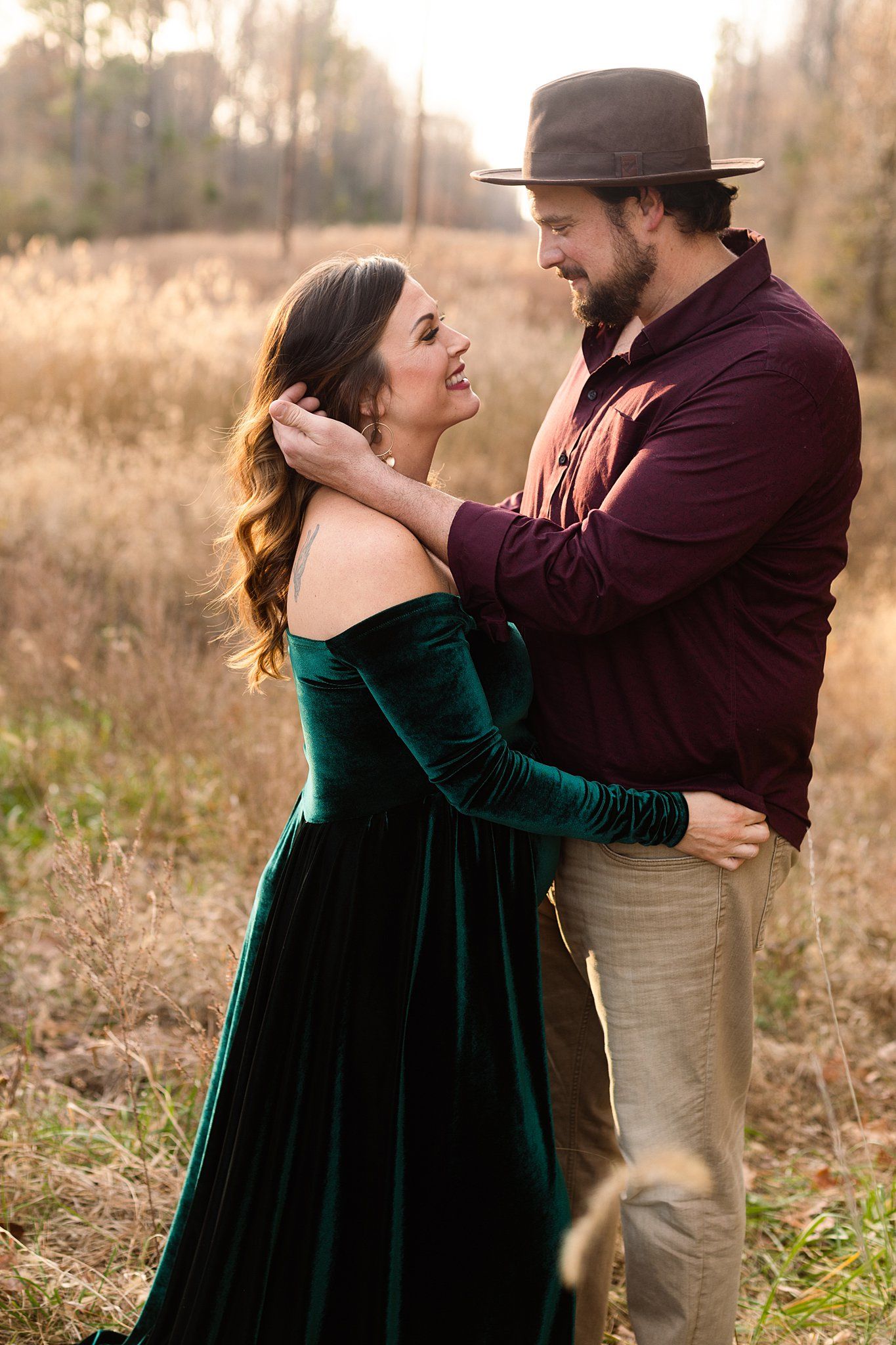 Intimate maternity photoshoot in a field with a couple embracing, woman wearing emerald velvet gown at sunset.