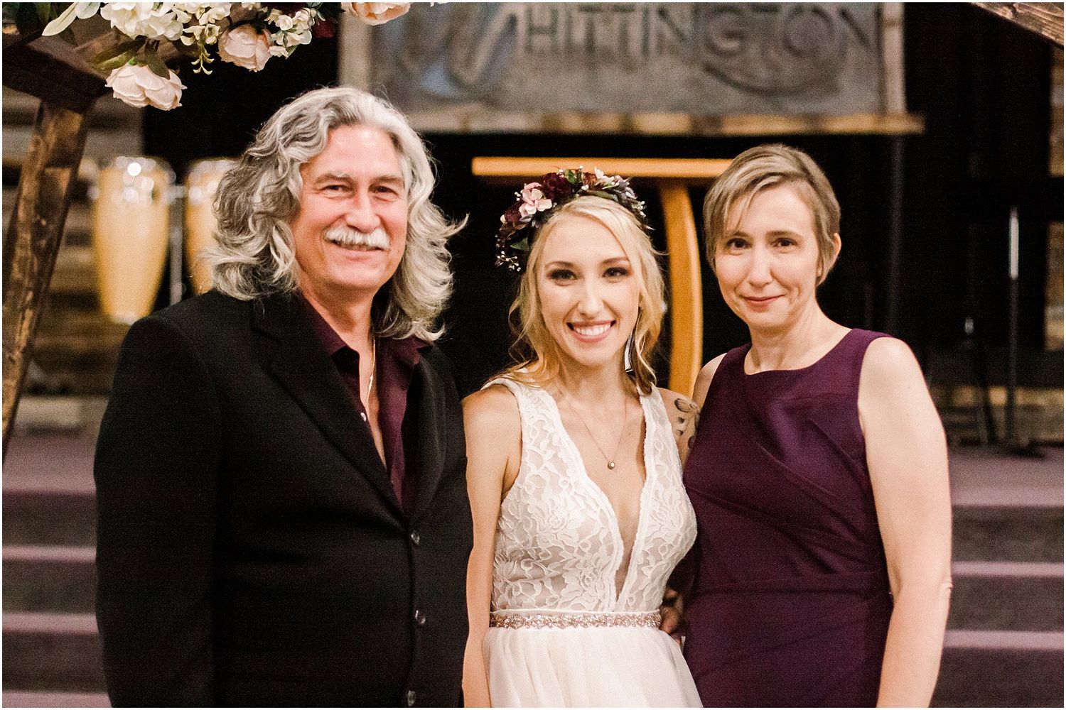 Three elegantly dressed people pose together at a rustic wedding venue.