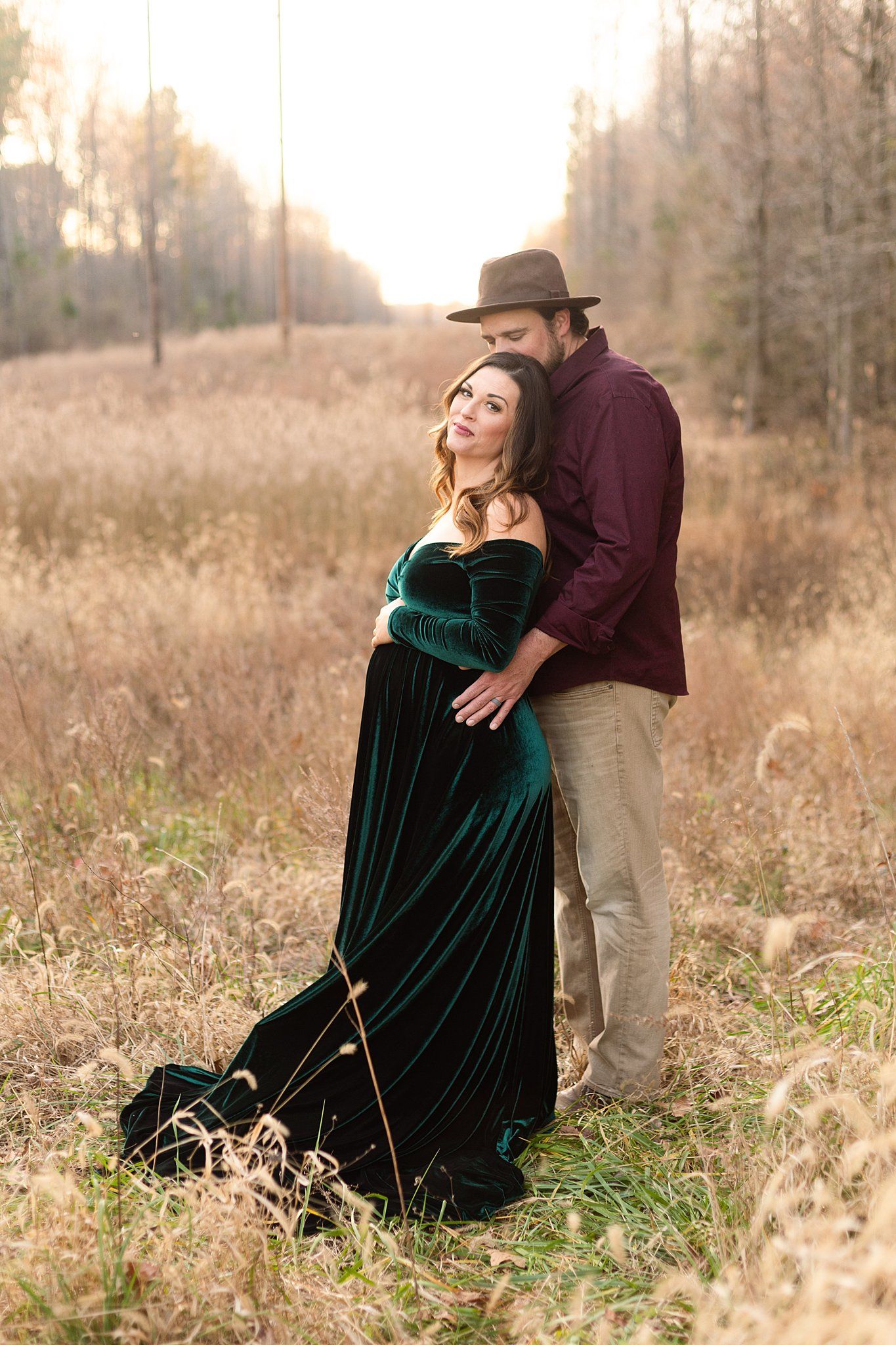 Dreamy autumn maternity photo with couple in burgundy shirt and flowing emerald dress at sunset.