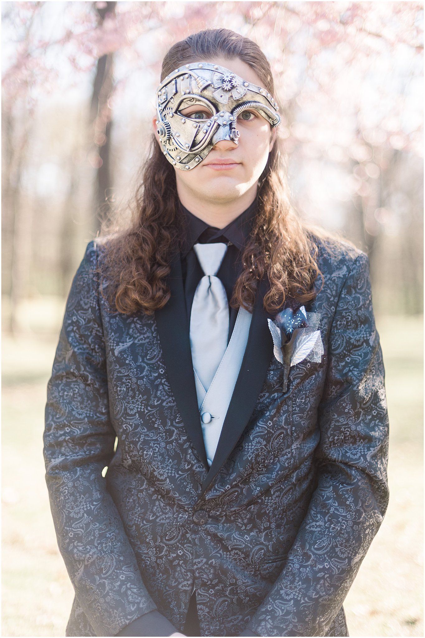 A person in a patterned blue suit and tie wearing an ornate silver masquerade mask.