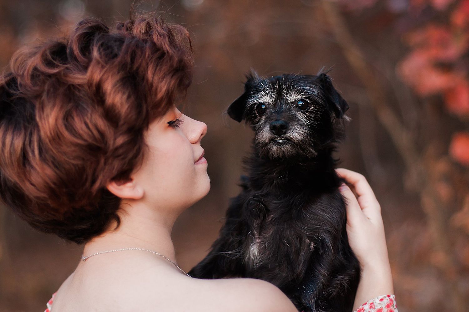 Person with short red hair holding a small black dog against autumn foliage.