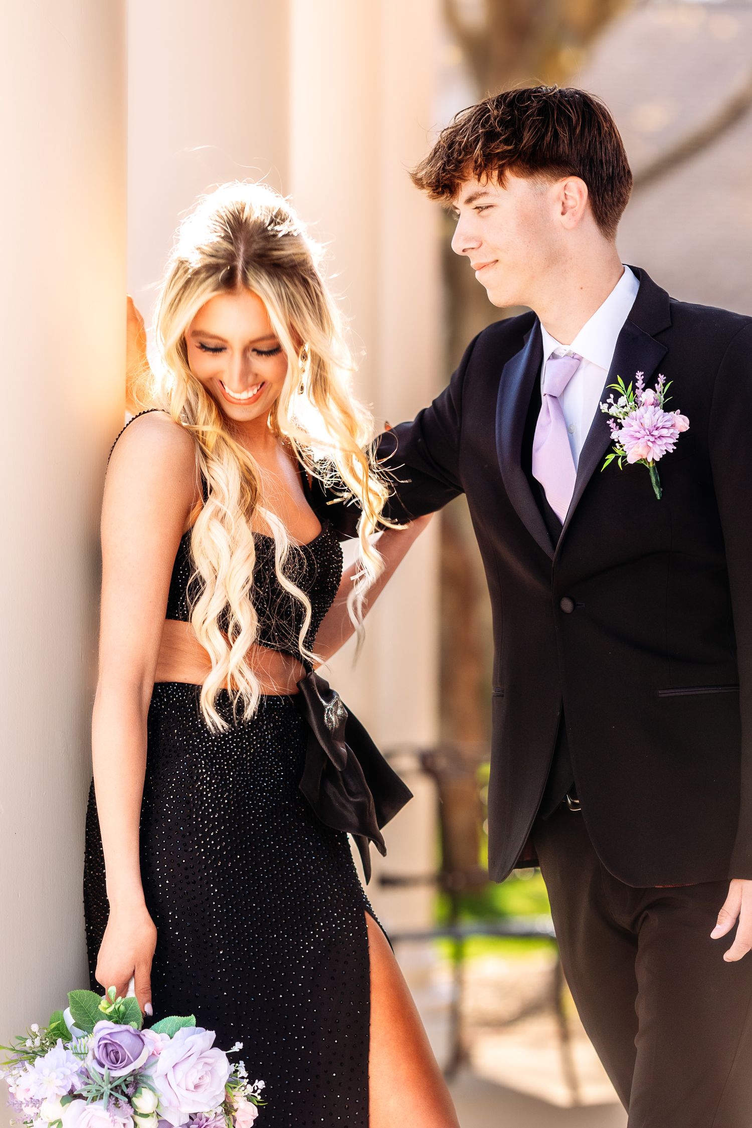 A couple in formal attire shares a romantic moment, with the woman wearing a black dress and holding purple flowers.