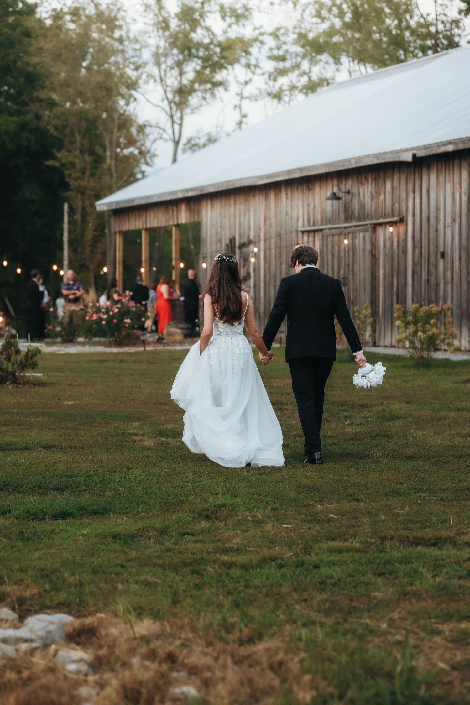 Newlyweds walk hand in hand towards a rustic barn venue with string lights at dusk.