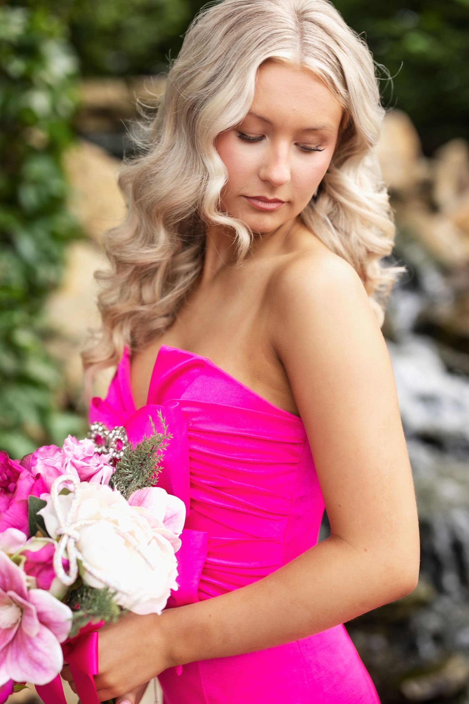 Blonde woman in pink strapless dress holding bouquet of pink and white flowers near waterfall.