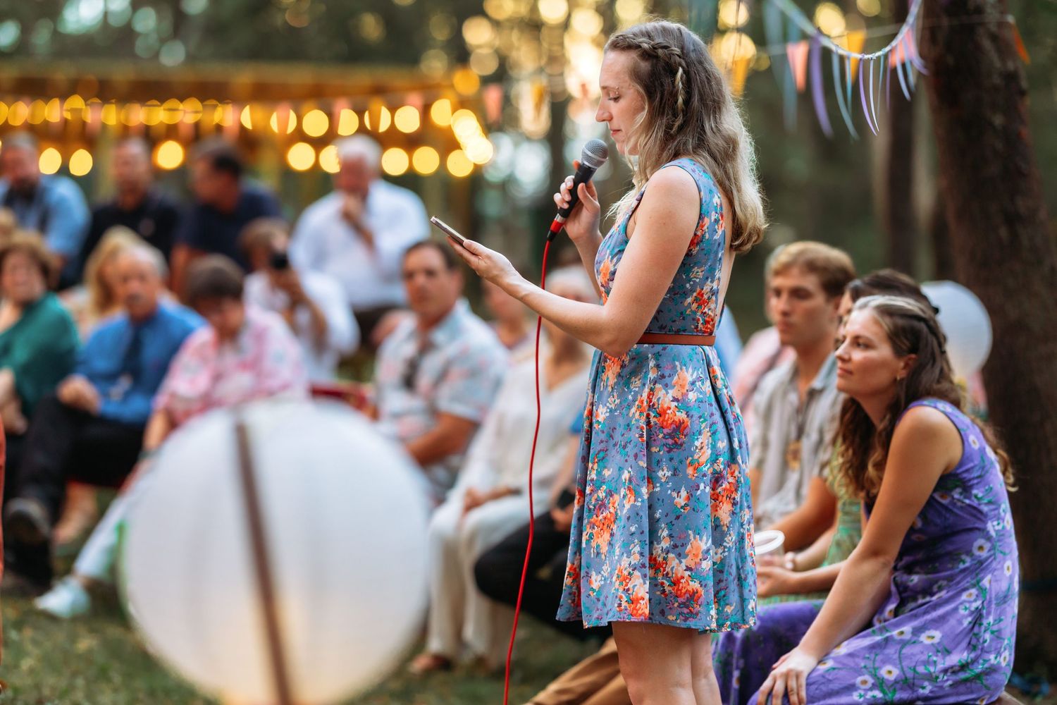 A guest delivers a speech at an outdoor wedding with string lights and seated guests in the background.