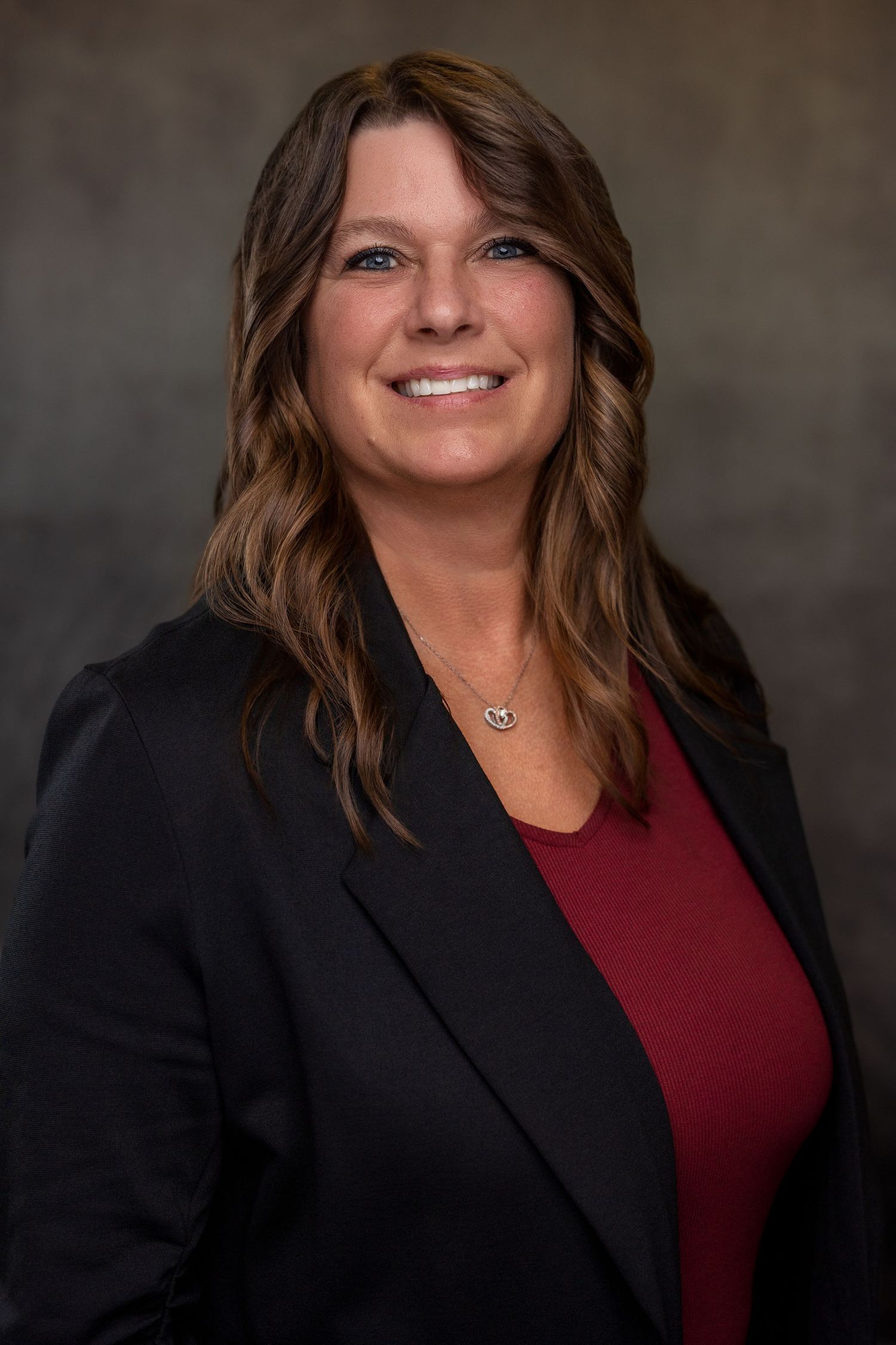 Professional portrait of woman in black blazer and burgundy top against neutral backdrop.