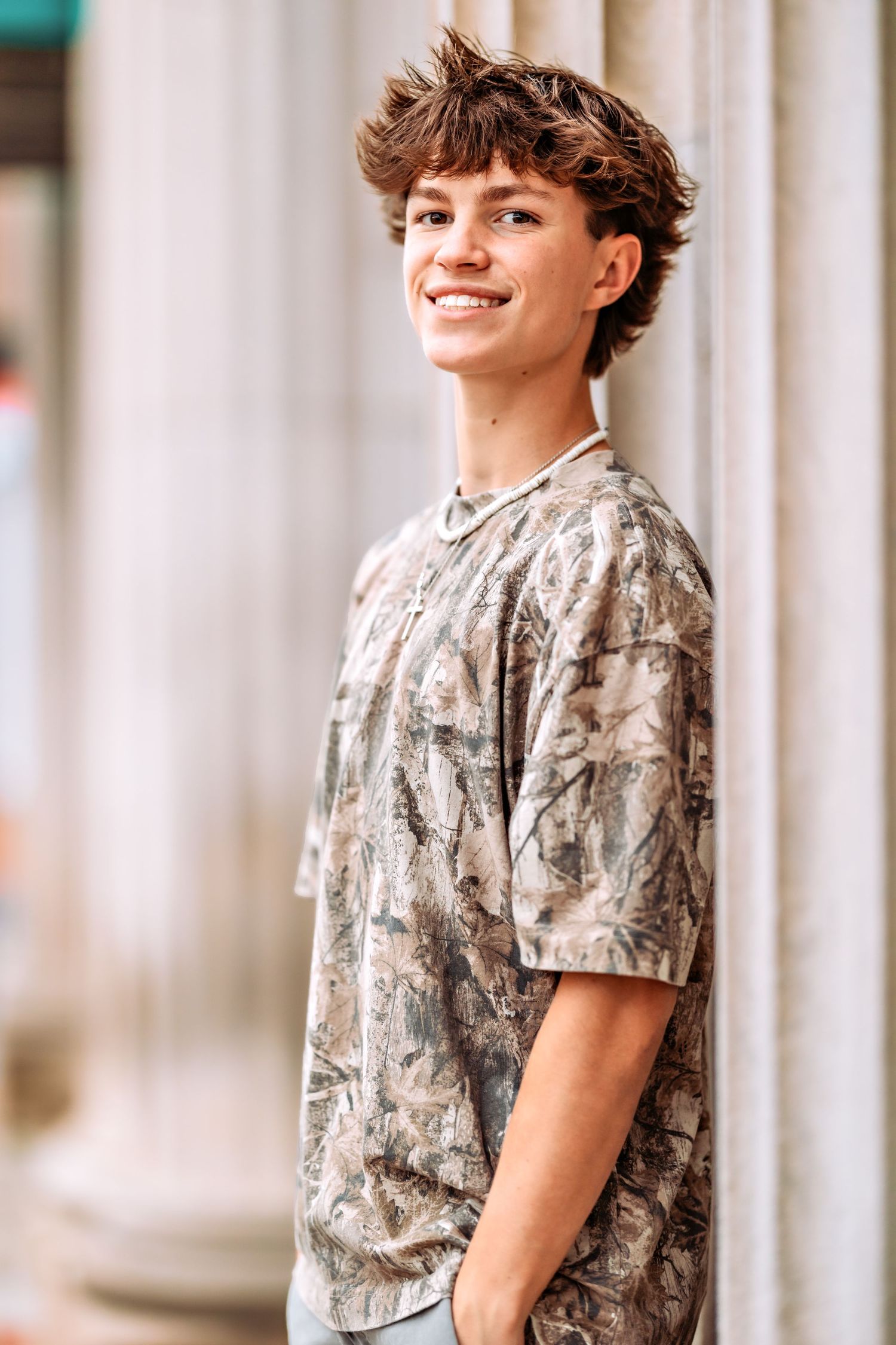 A young person in a camouflage t-shirt poses against white columns with a casual smile and tousled brown hair.