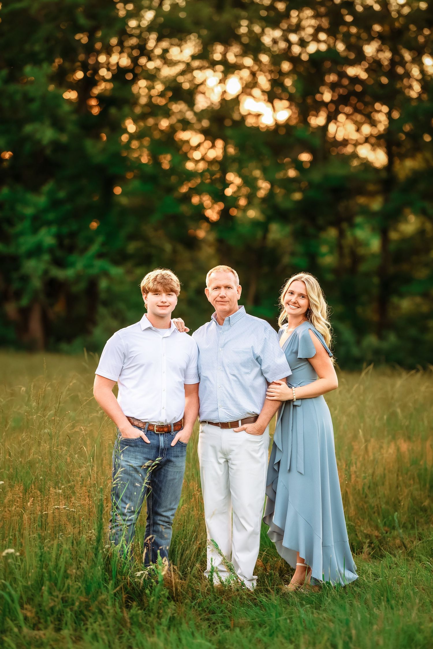 A family wearing coordinated blue and white outfits poses together in a grassy field at golden hour.