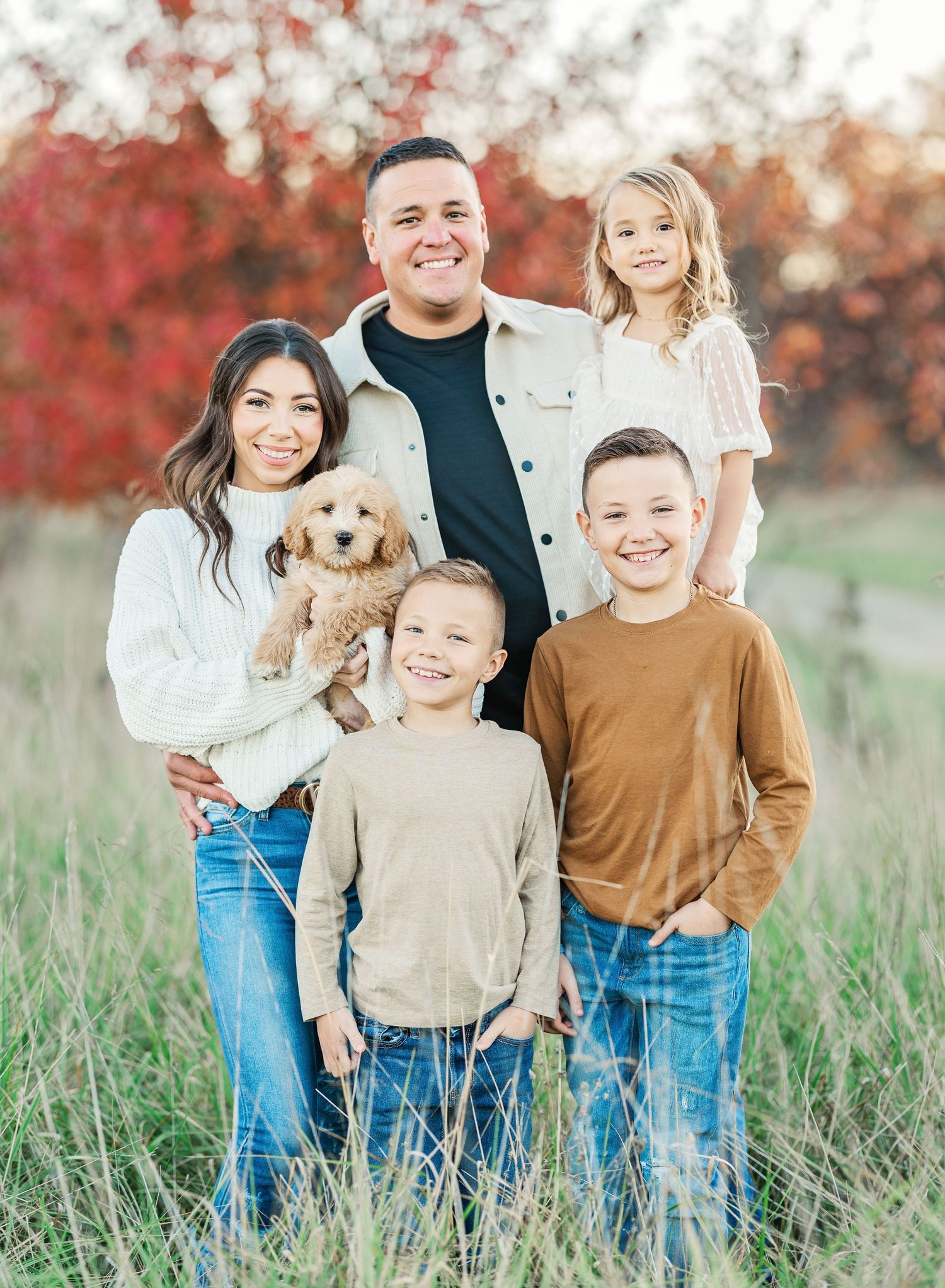 Family with puppy poses together in field with red autumn foliage in background.