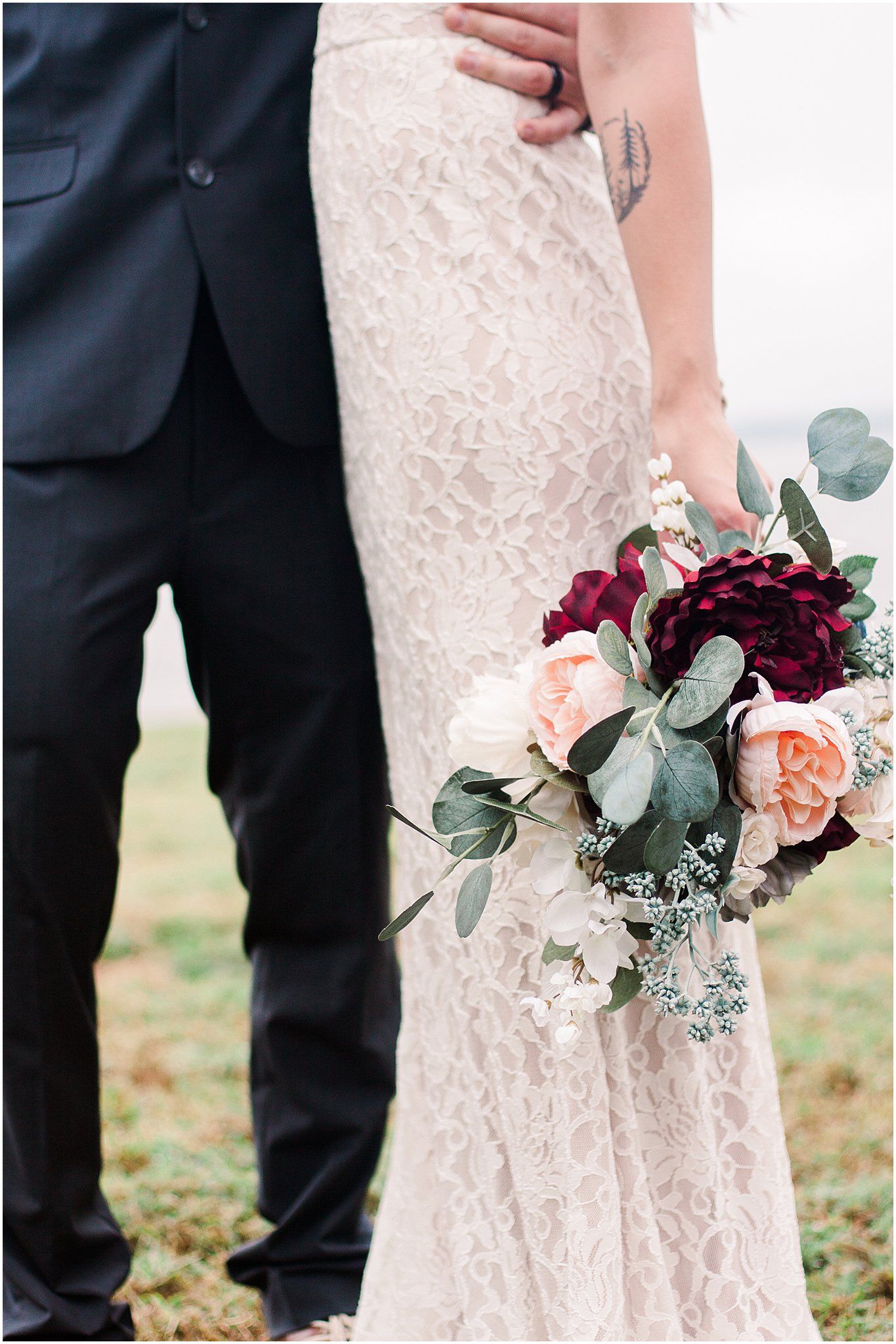 Close up of wedding bouquet with burgundy and pink roses against lace dress and dark suit.
