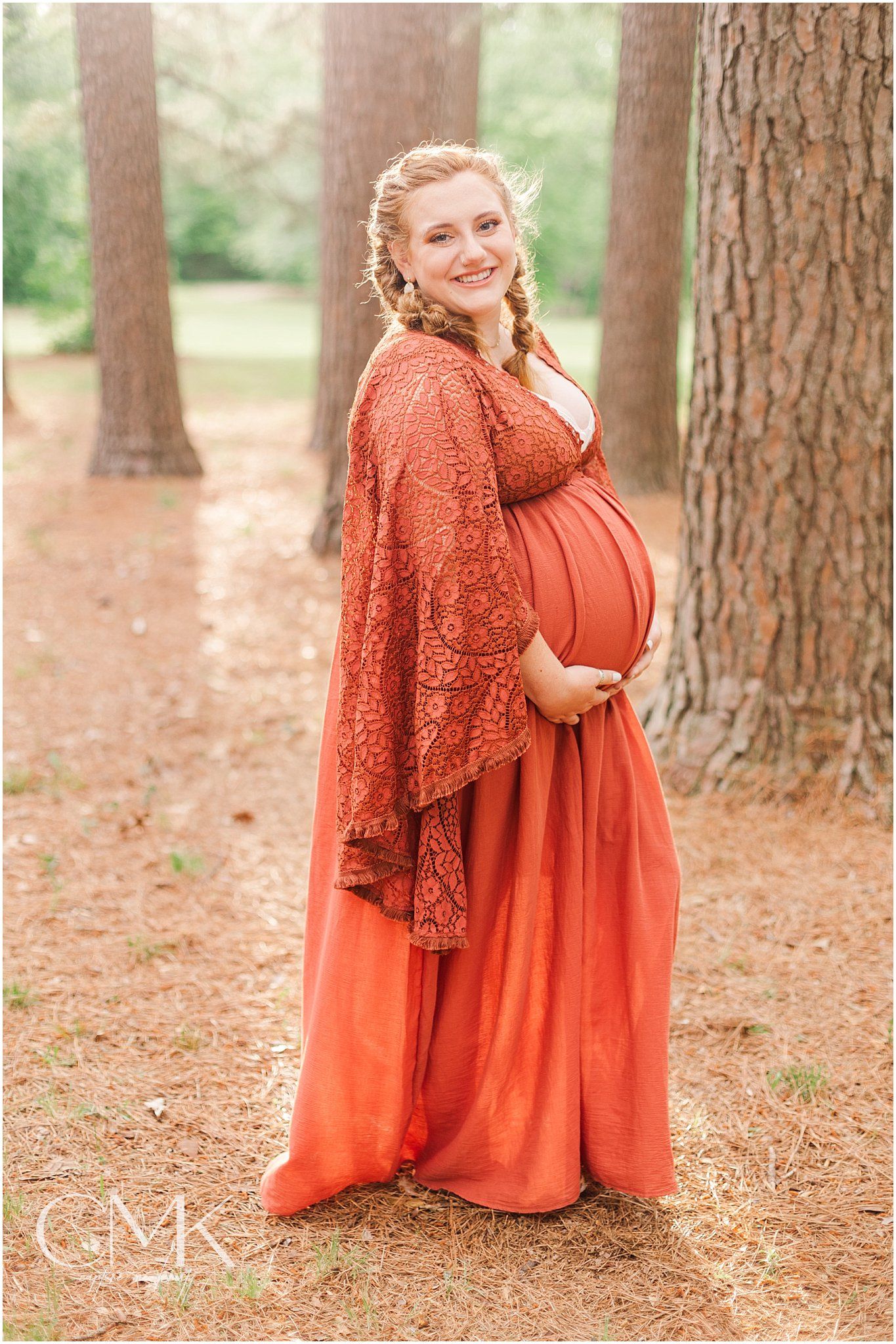 Expectant mother in a burnt orange maxi dress and lace shawl poses among pine trees.