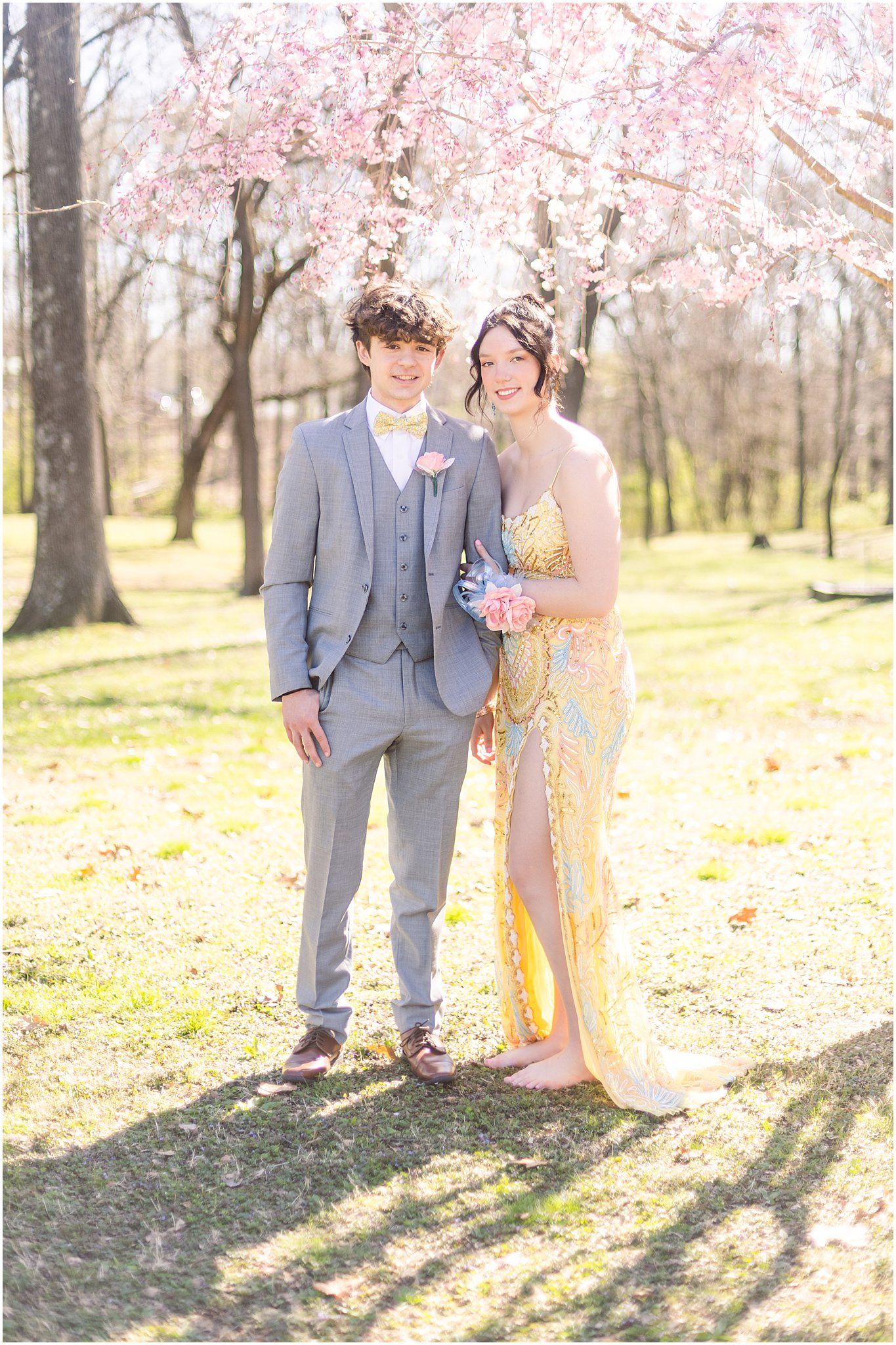 Formal portrait of couple in grey suit and yellow dress under cherry blossoms.