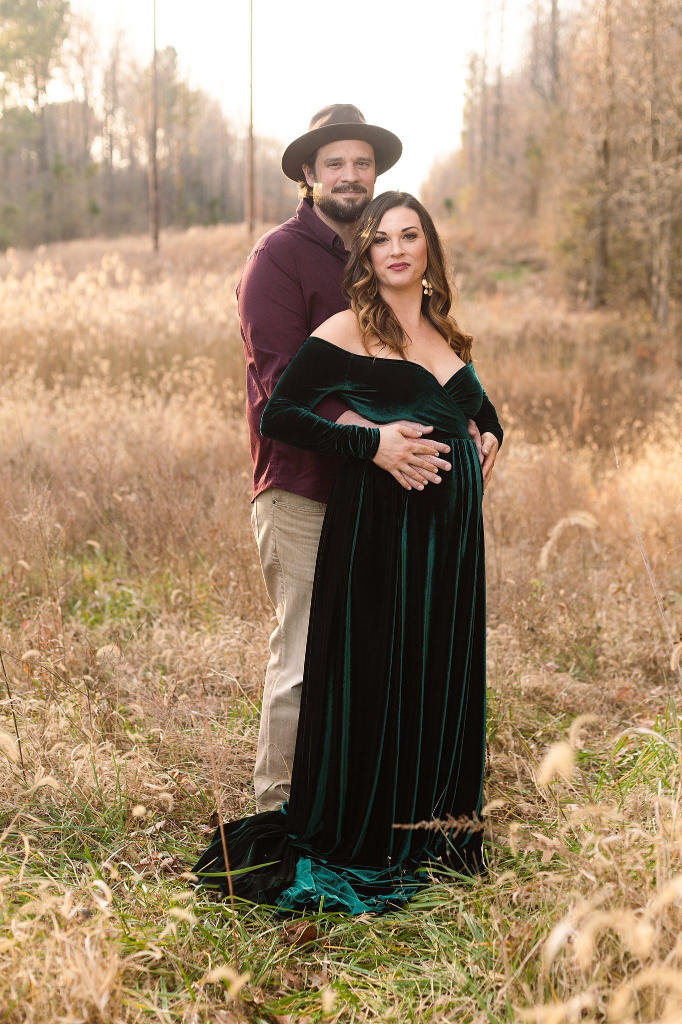 A couple poses in a field during autumn, wearing a burgundy shirt and emerald velvet dress.