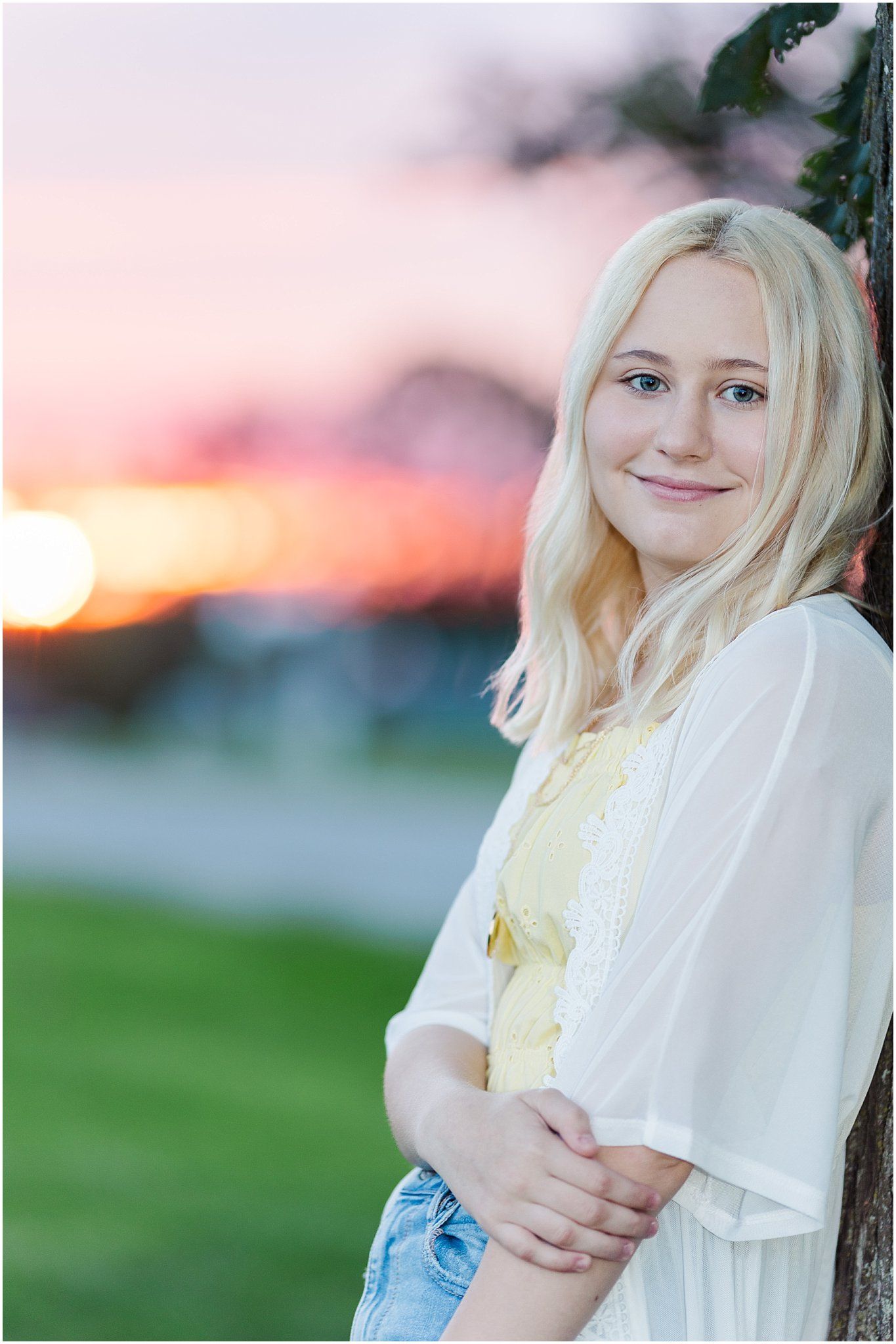 Senior girl leaning against a tree by a corn field with a pink and orange sunset in the background
