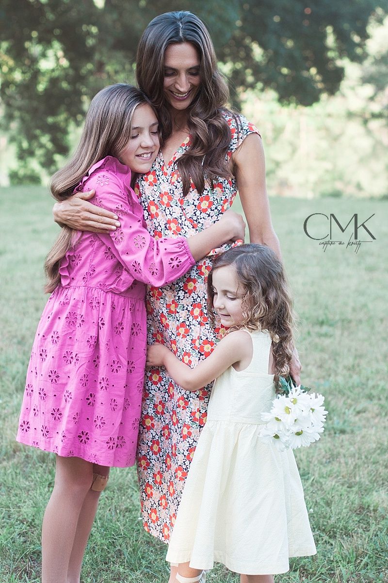 Family portrait showing three people hugging and smiling in colorful summer dresses outdoors.