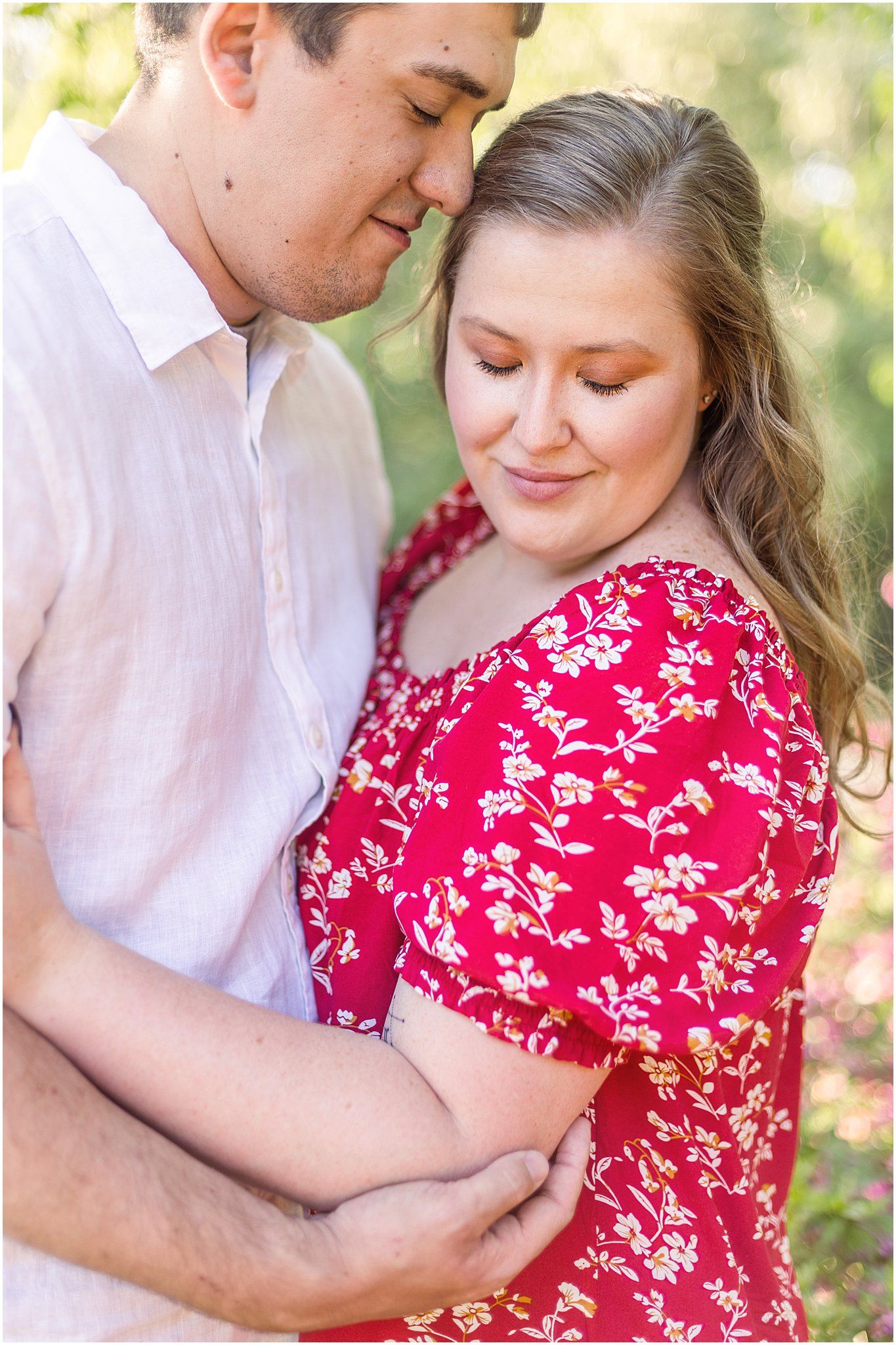 Tender embrace captured in soft lighting as couple in white and red floral dress pose together outdoors.
