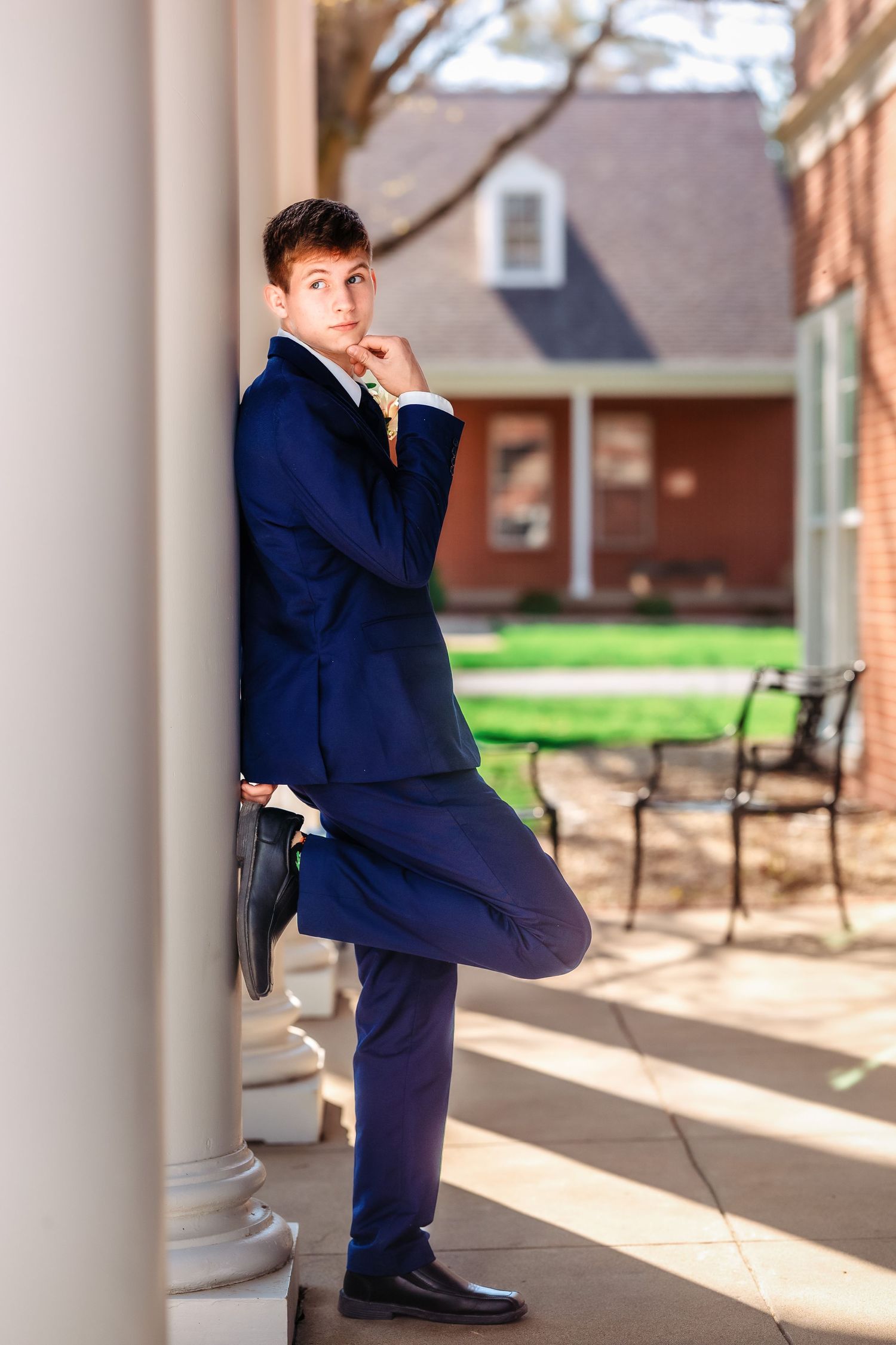 A person in a navy suit strikes a playful pose while leaning against a white column on a brick building's porch.
