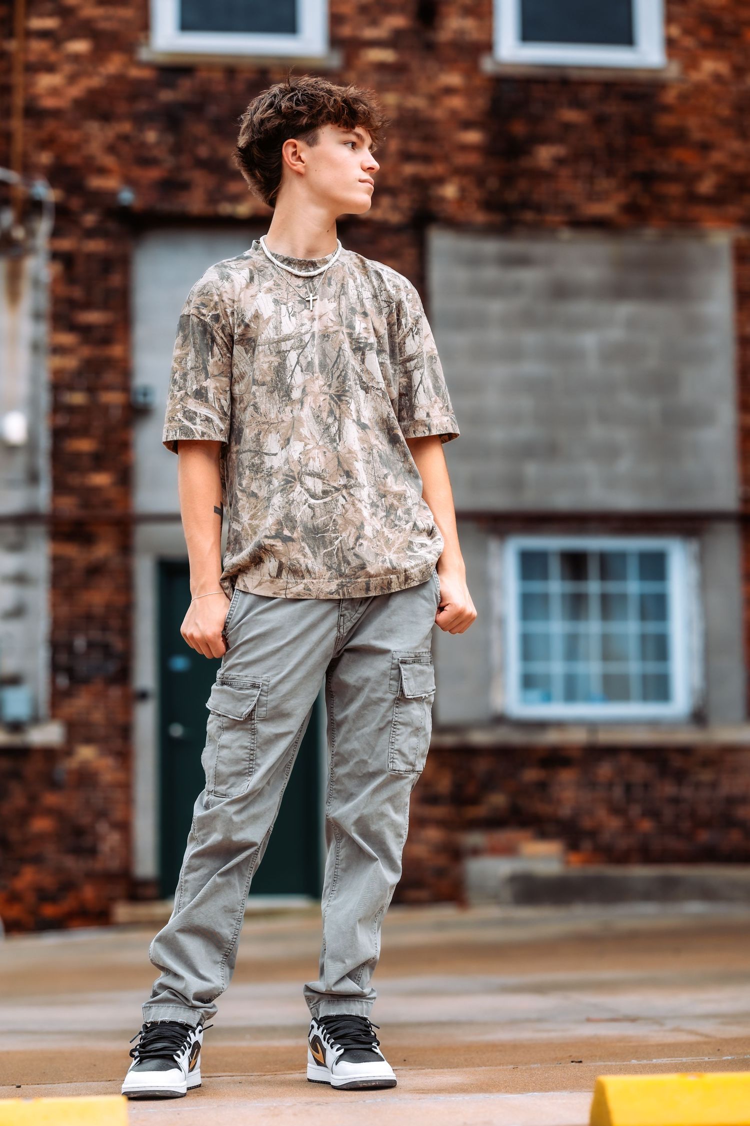 Urban fashion shot featuring camouflage t-shirt and gray cargo pants against brick building backdrop.