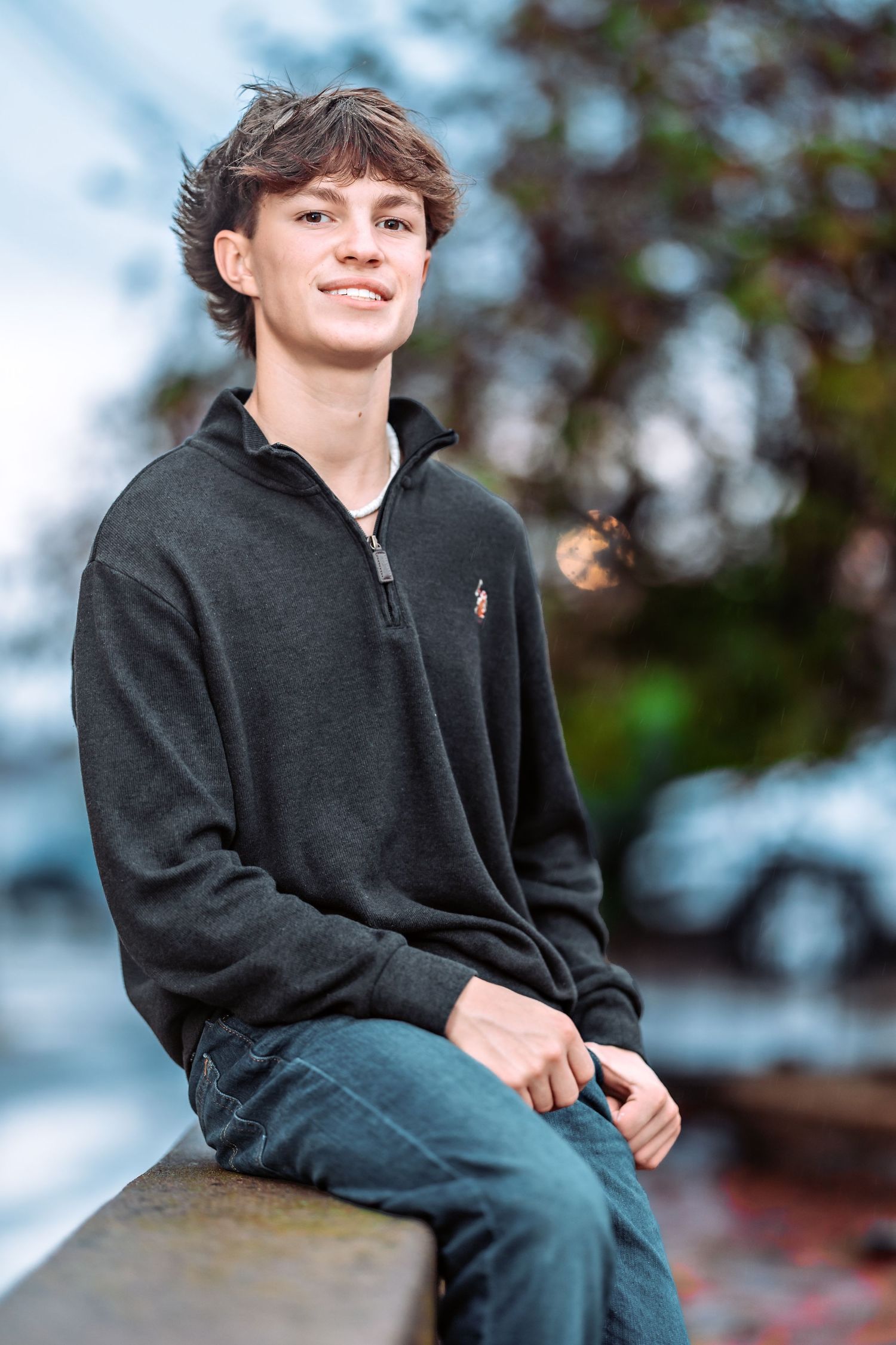 A young person in a black quarter-zip sweater sits casually outdoors on a sunny day with trees in the background.
