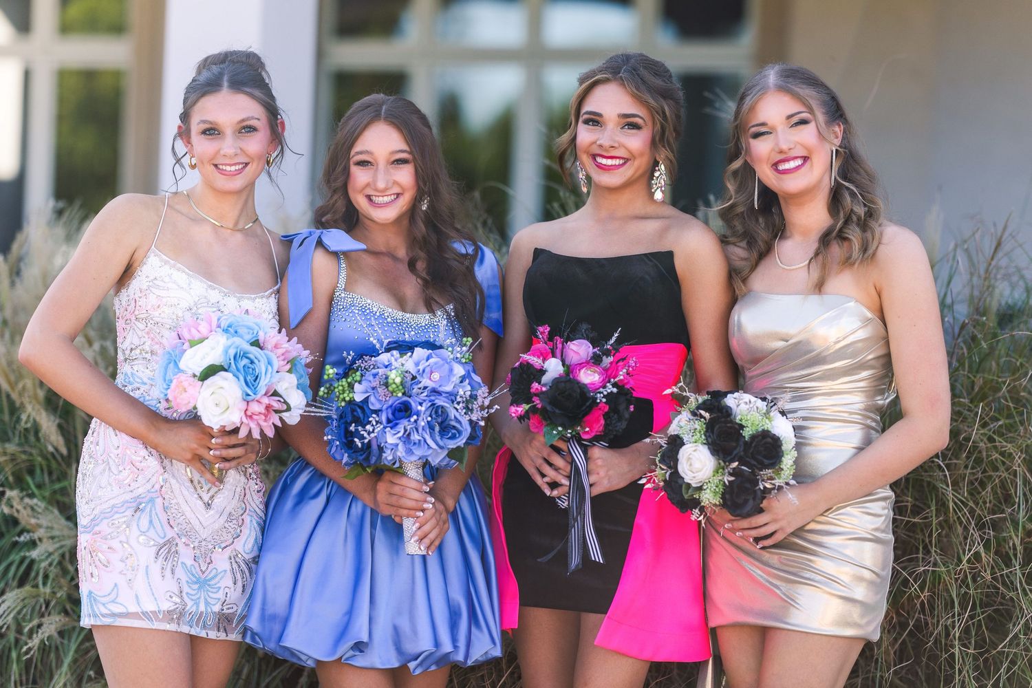 Four friends in colorful cocktail dresses pose together holding floral bouquets.