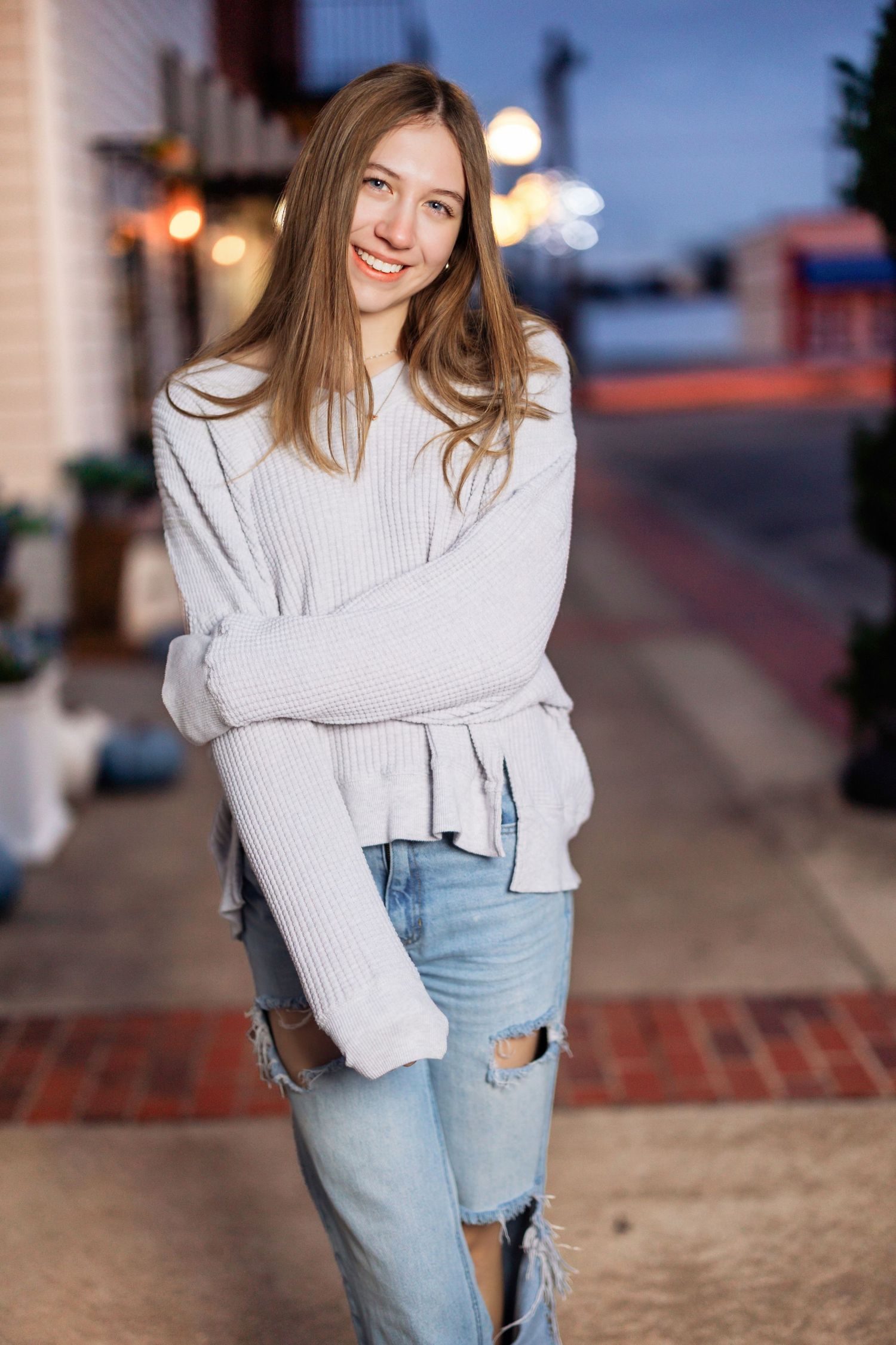 Person in cozy white sweater and distressed jeans poses on city street at dusk.