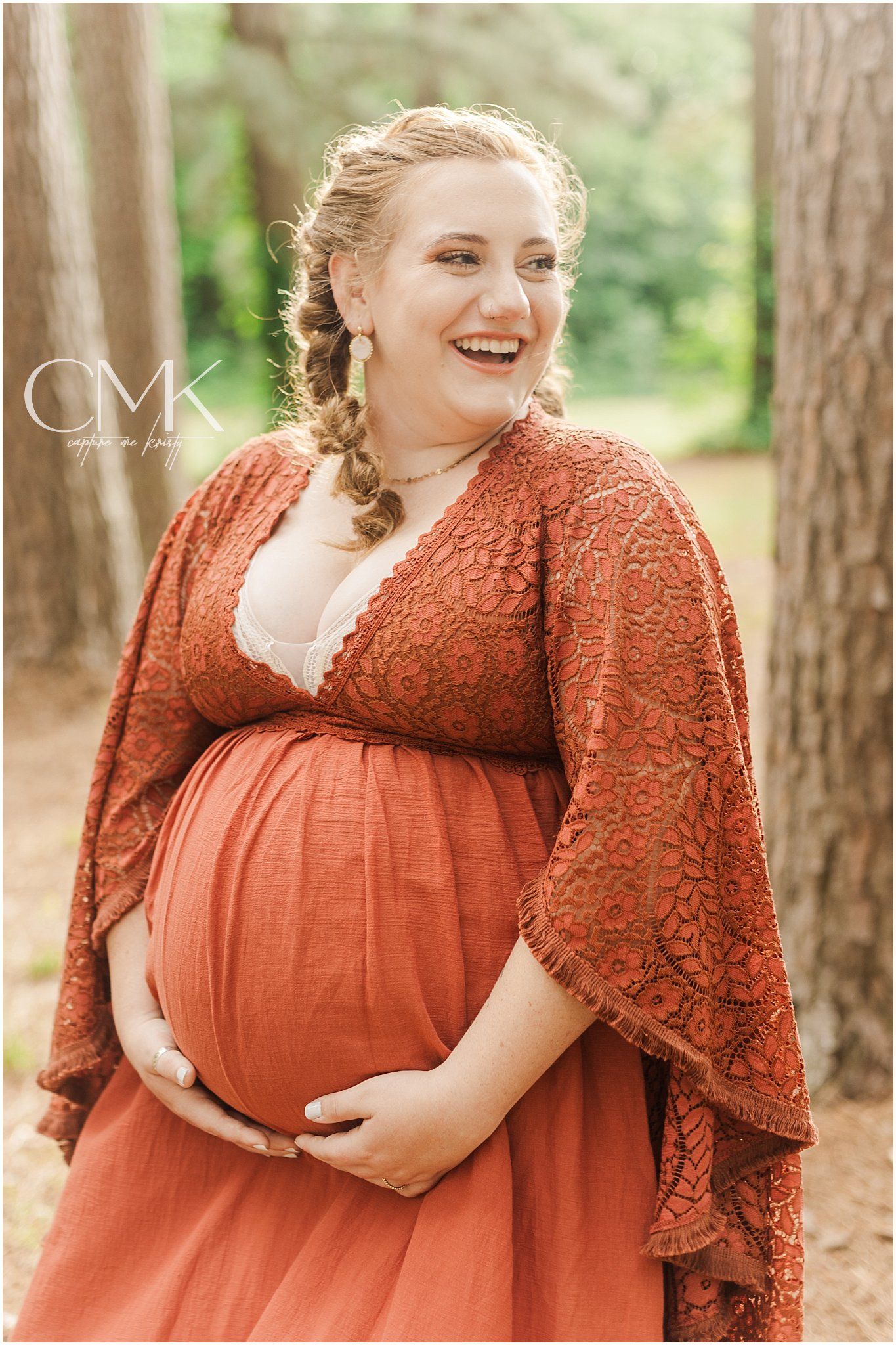 Expectant mother wearing a flowing rust-colored dress poses between tall pine trees.