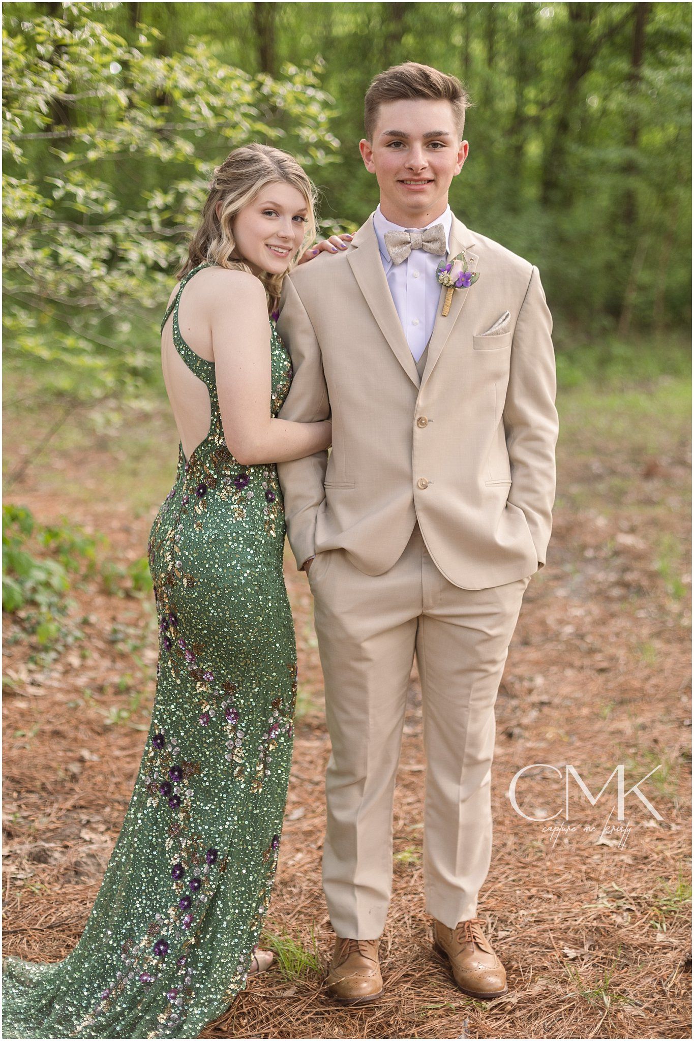 A formal portrait in the woods featuring a sparkling green dress and beige tuxedo with bow tie.