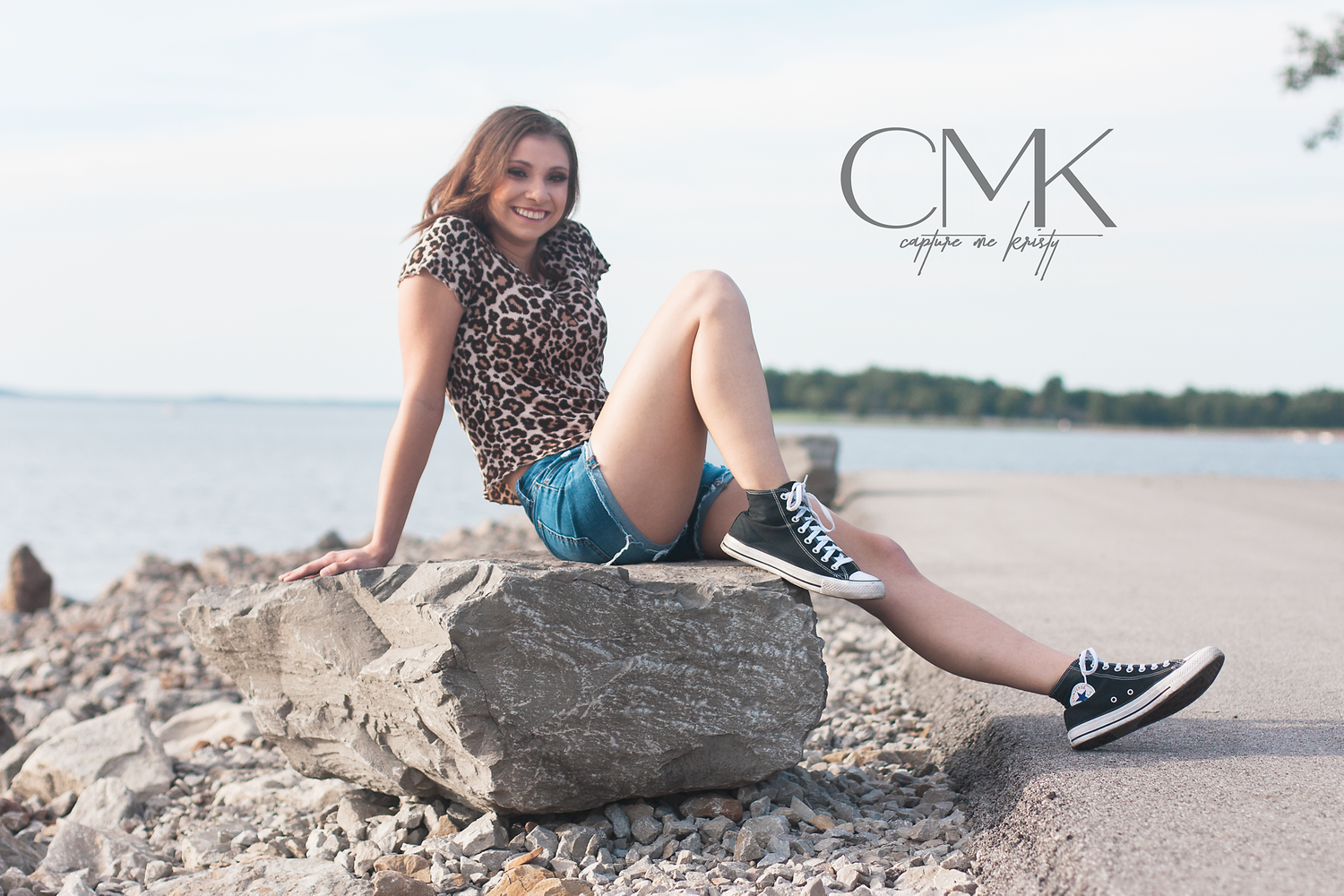 Portrait of someone in leopard print top and denim shorts sitting casually on large rock by waterfront.