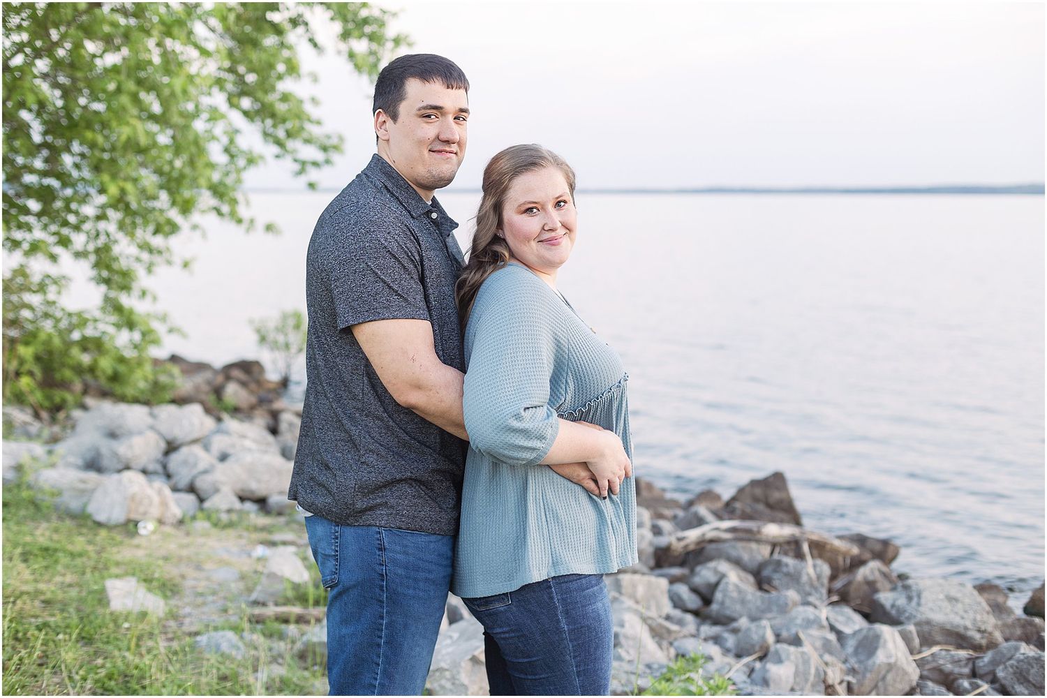 A couple embraces while posing by a rocky lakeshore on a cloudy day.