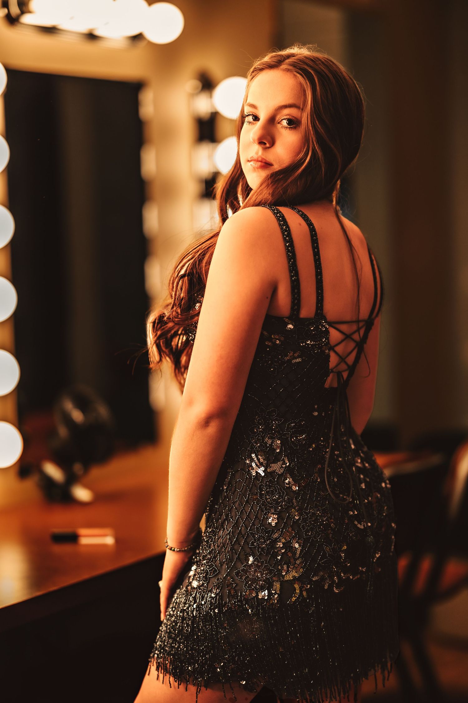 Person wearing elegant black sequined dress poses in dimly lit room with bokeh lights.