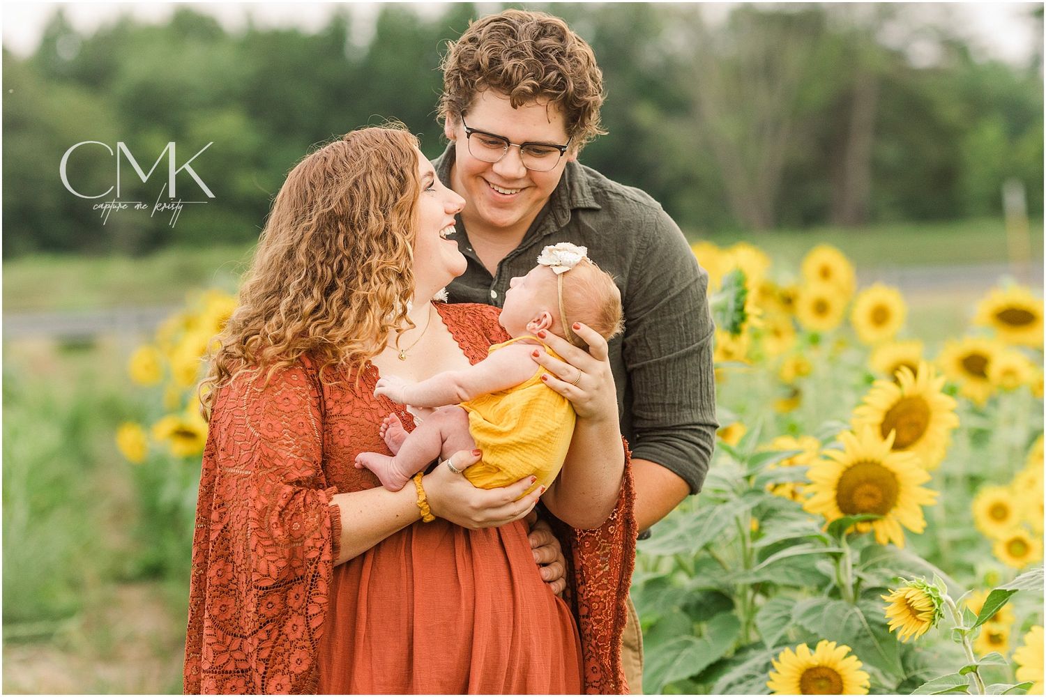 Family shares tender moment together in vibrant sunflower field while wearing rust-colored dress and olive shirt.